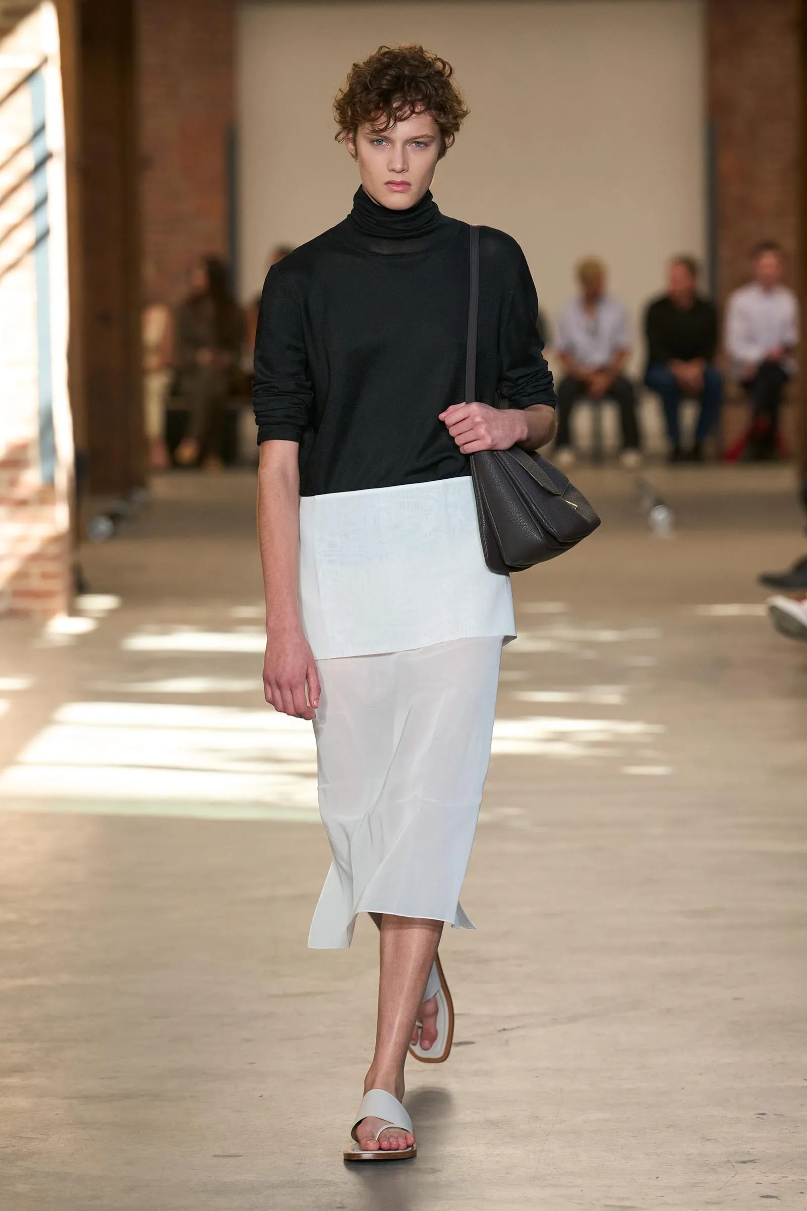 Model walks a runway wearing a black turtleneck and white layered skirt, carrying a gray shoulder bag and white sandals.