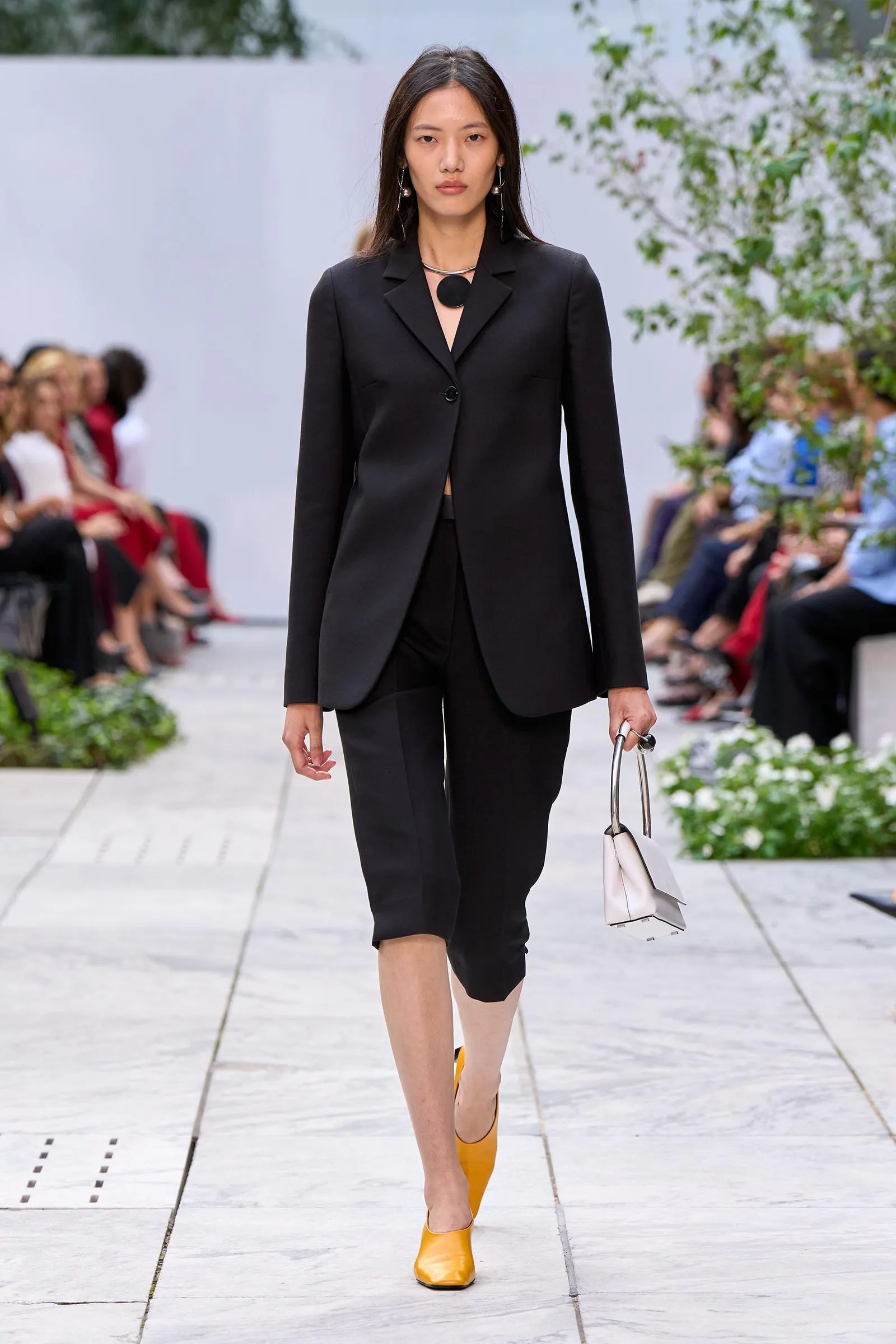 Model walks a runway in a black blazer and matching pants, bright yellow shoes, and a light handbag on her right.