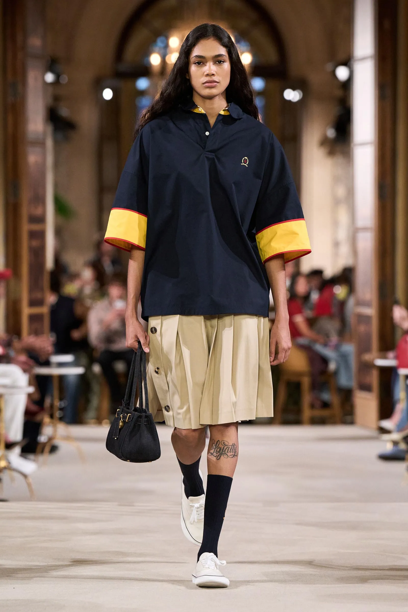 Model walking the runway in a navy top with yellow-cuffed sleeves and a beige pleated skirt, carrying a black handbag.