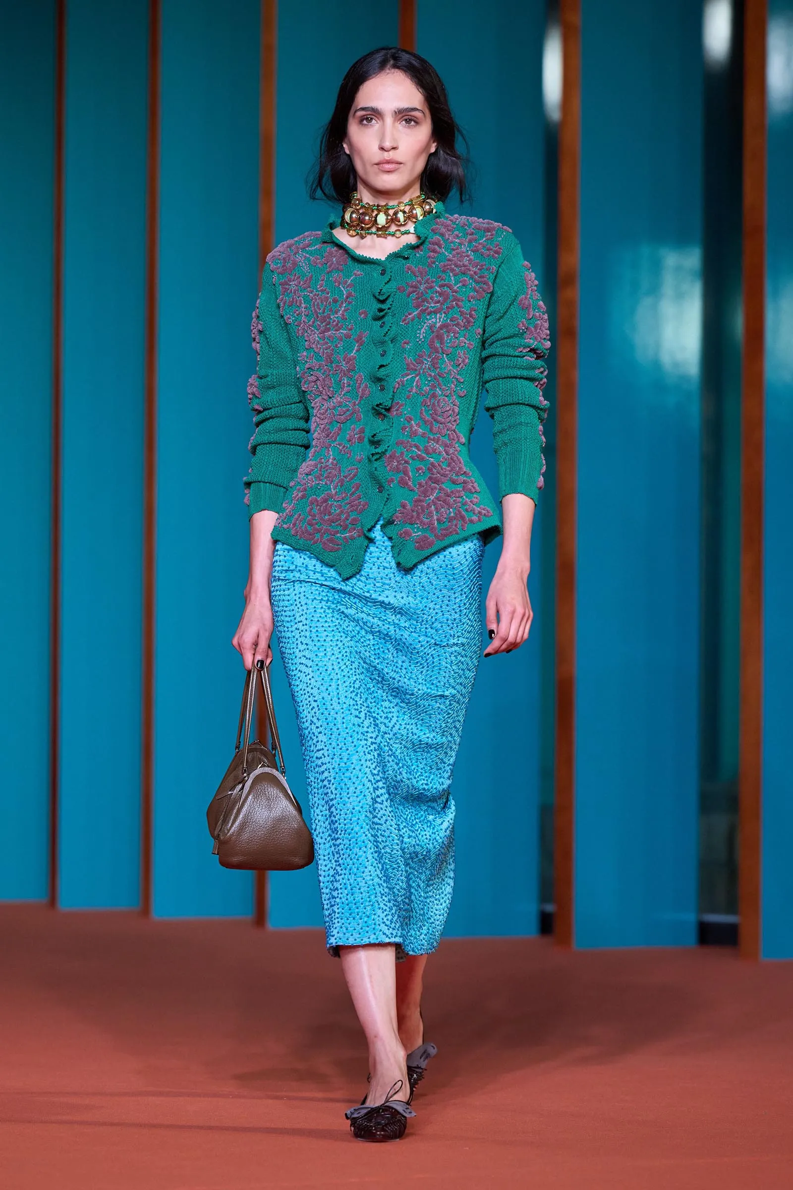 Model walking runway in teal cardigan with purple floral embroidery, turquoise patterned skirt, and chunky green bead necklace, carrying a brown handbag.