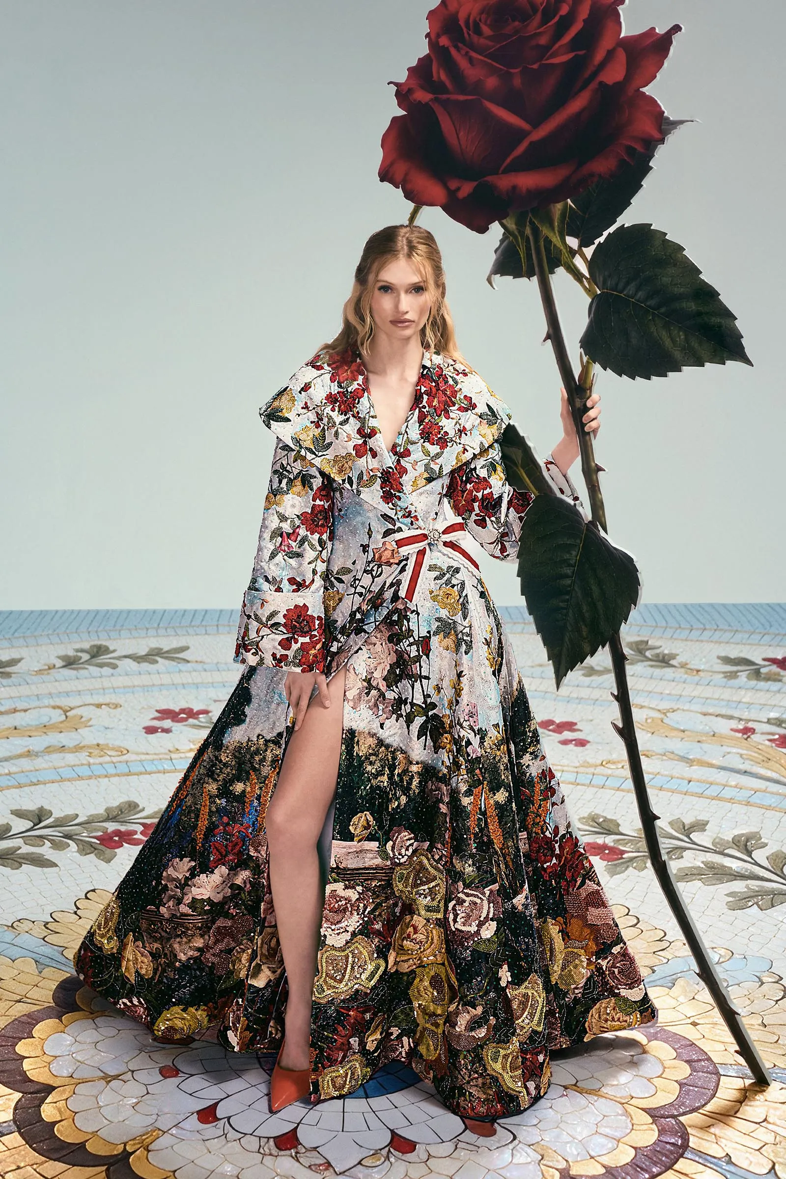 Model in a richly embroidered floral gown poses with a giant red rose prop indoors, standing on a decorative mosaic floor.