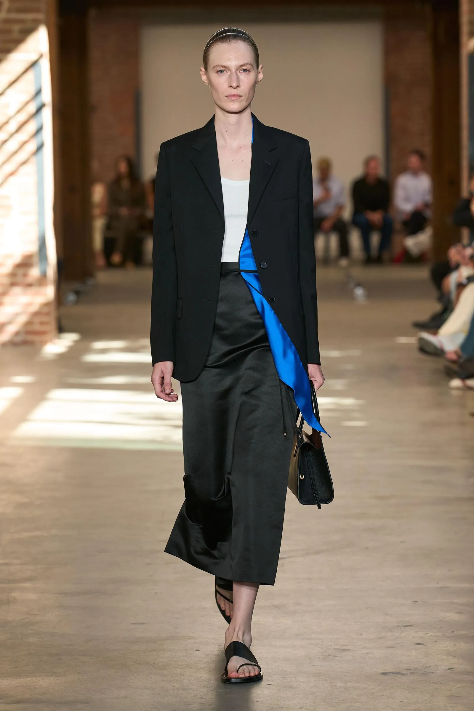 Model walks down a runway in a black blazer over a white top with a bright blue sash, black skirt, and black sandals, in a studio-like venue with blurred audience in the background.
