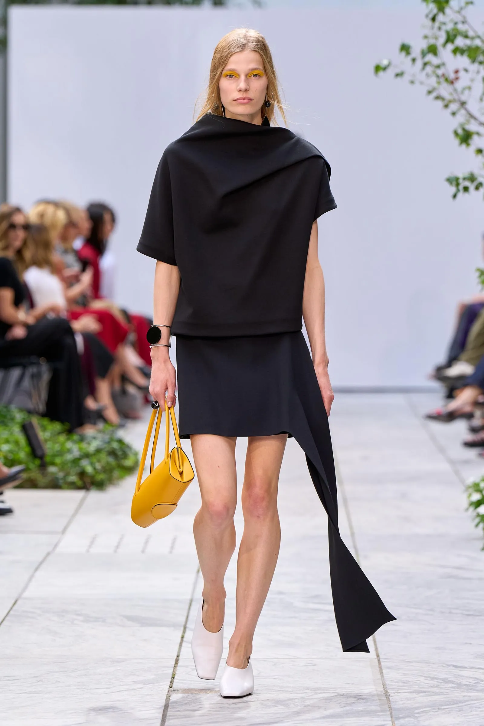 Model walks a fashion runway in a black draped top with an asymmetric hem, carrying a bright yellow handbag; blurred audience in the background.