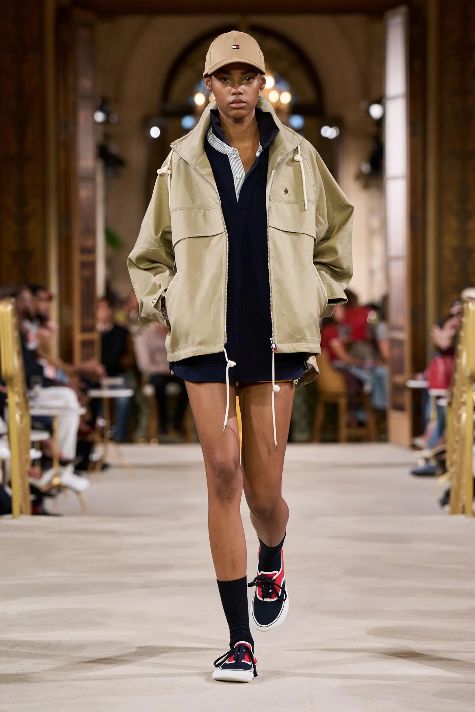 Model walking the runway in a beige cap, oversized beige jacket, navy shirt and navy shorts with red sneakers.