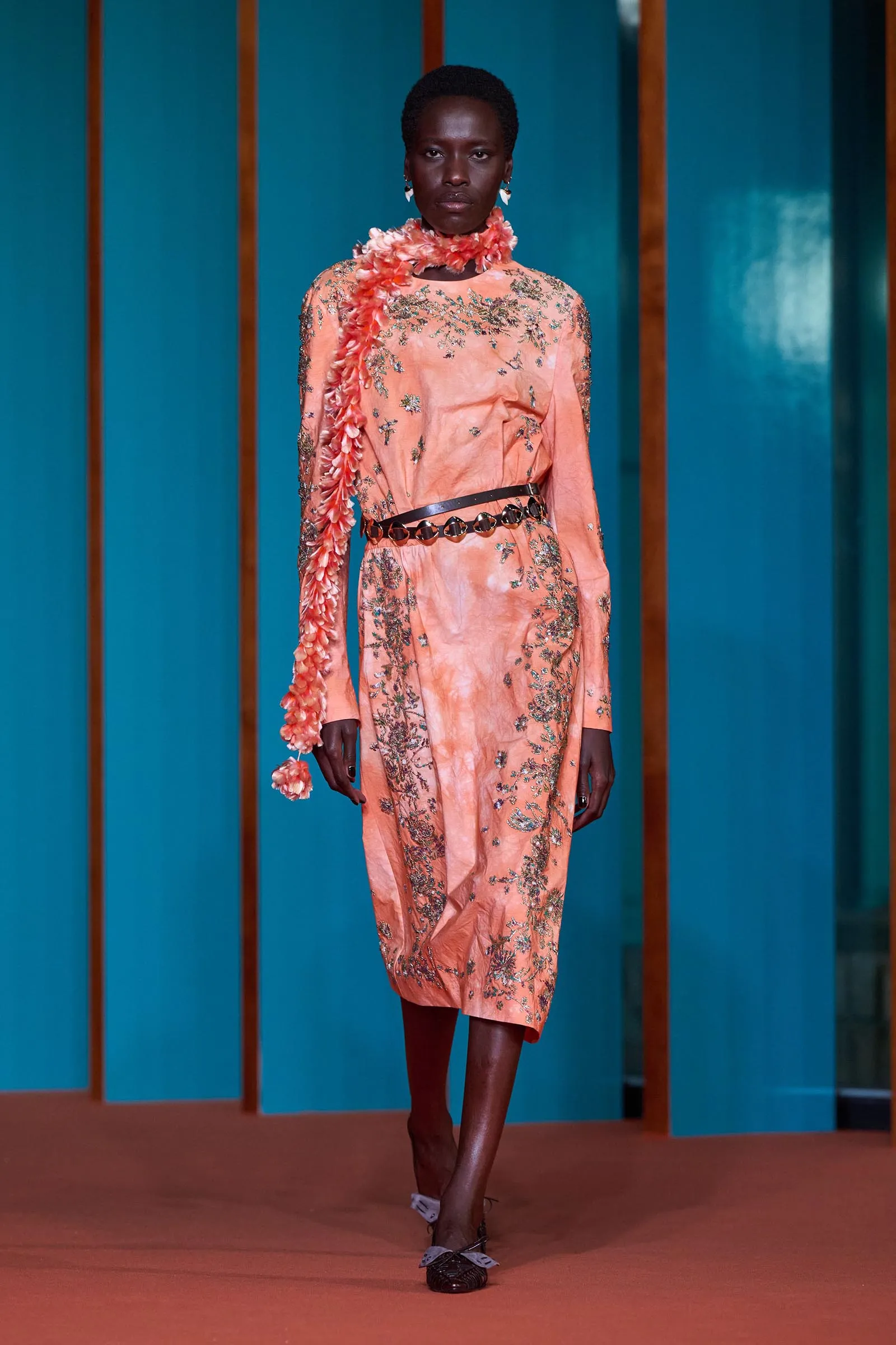 Model walking a runway in a peach embroidered gown with a floral lei scarf draped around the neck on a teal backdrop.
