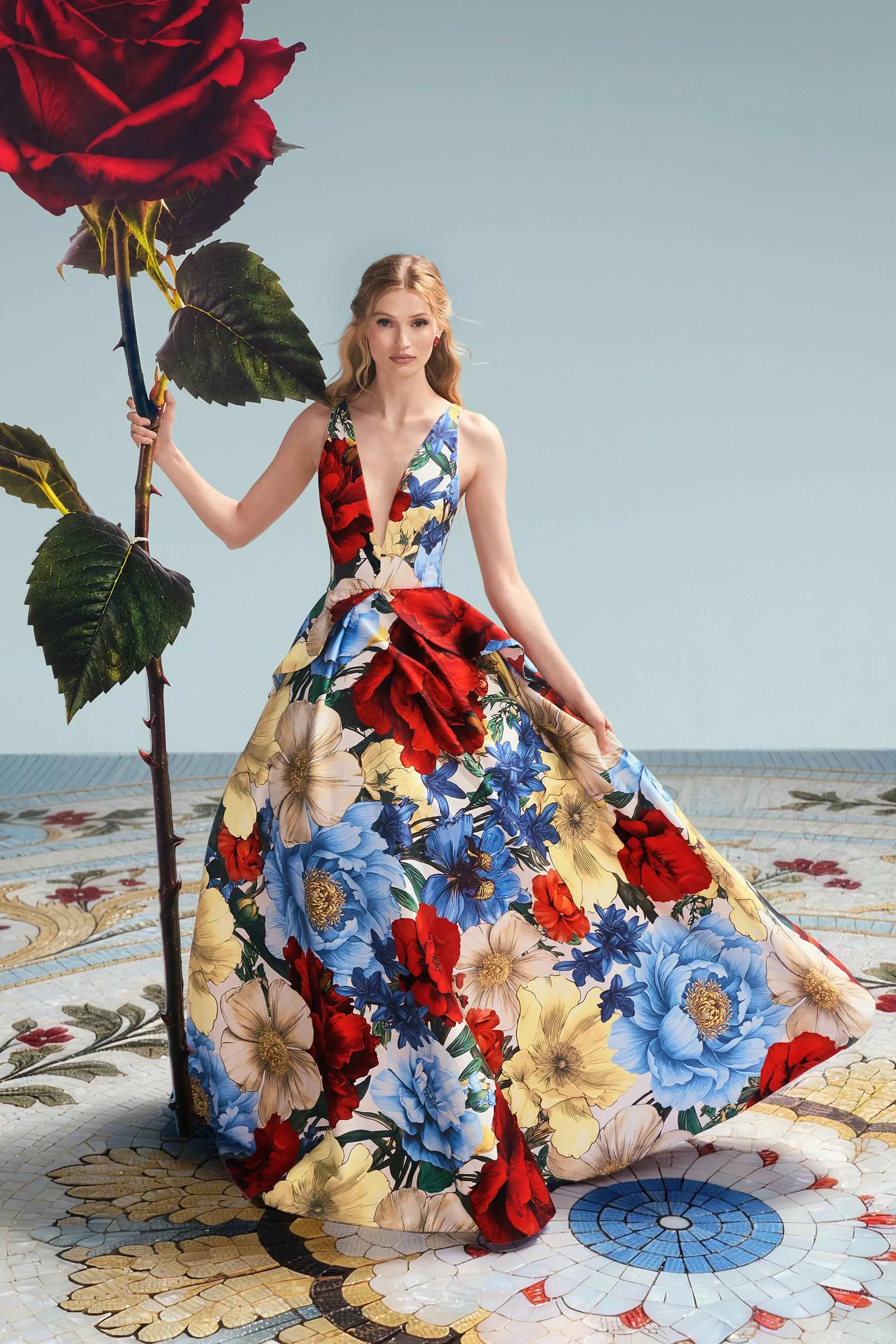 Woman in a bold, multicolored floral dress holds a tall red rose prop beside a patterned floor.