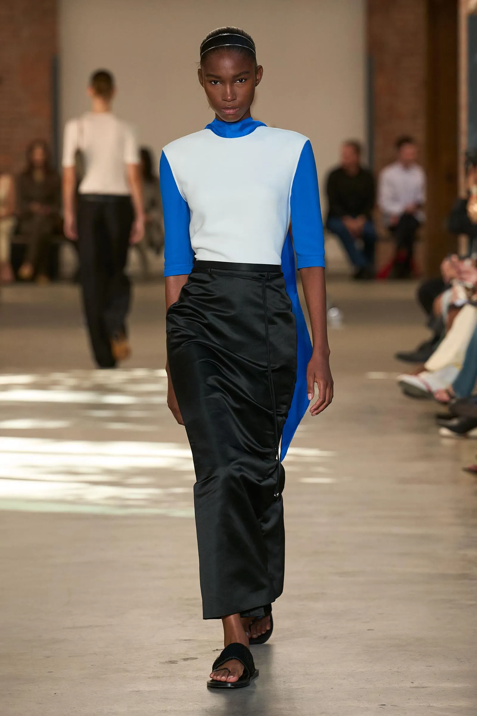 Model walks down runway wearing a blue long-sleeve top with a light sleeveless white overlay and black skirt, black headband visible in hair.