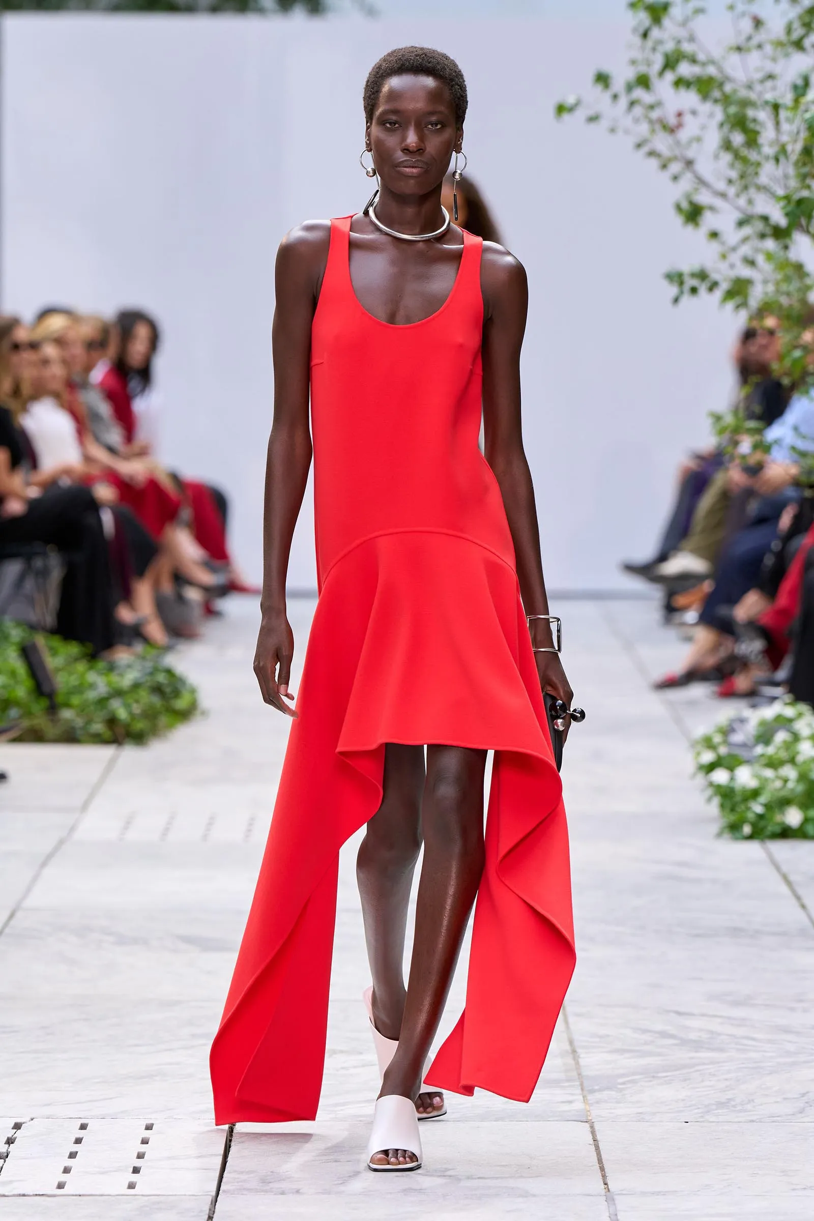 Model walks the runway in a bright red sleeveless high-low dress with silver jewelry during a fashion show.