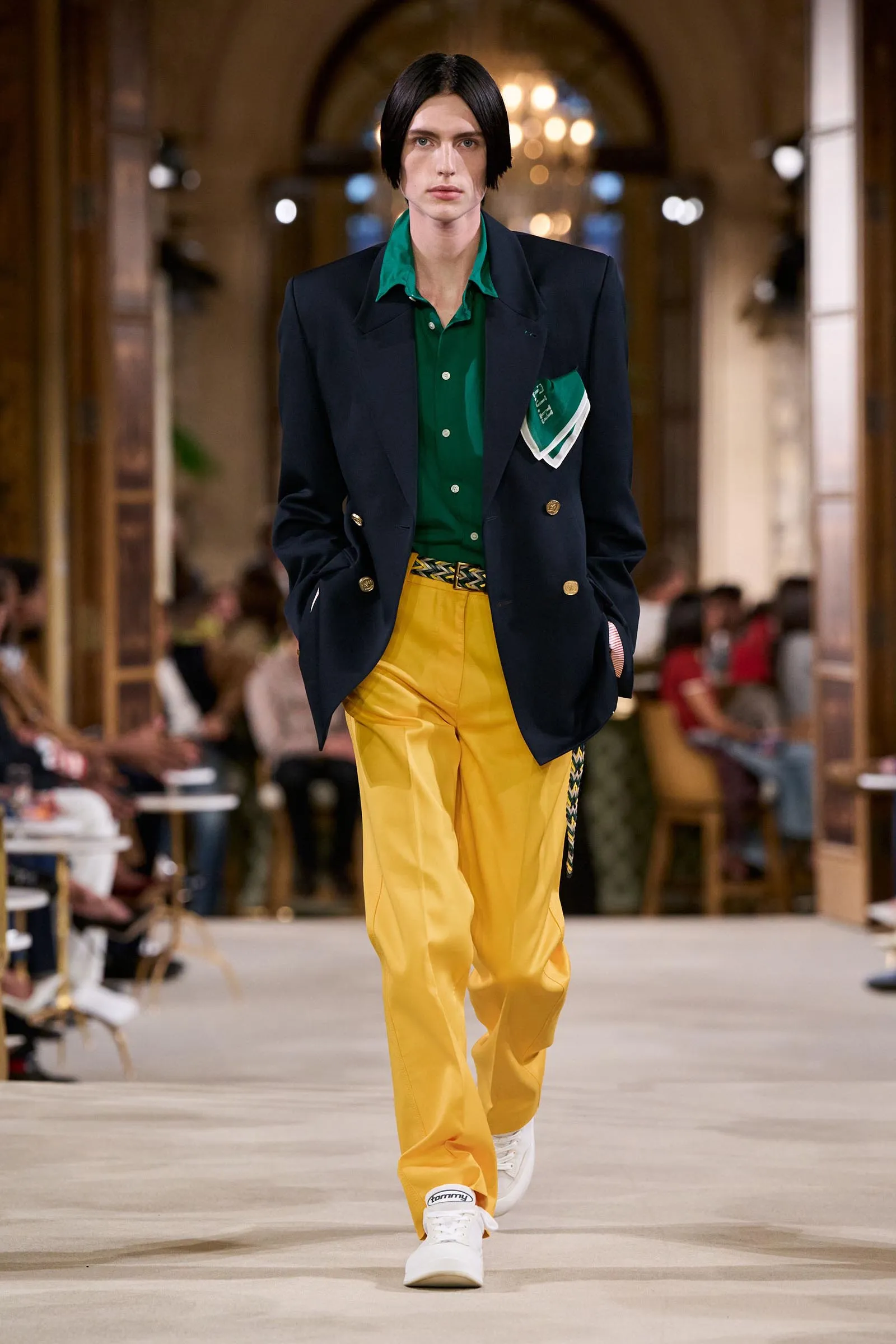 Model walks a fashion runway in a navy blazer, green shirt, and yellow pants; white sneakers, indoor venue with seated audience in the background.