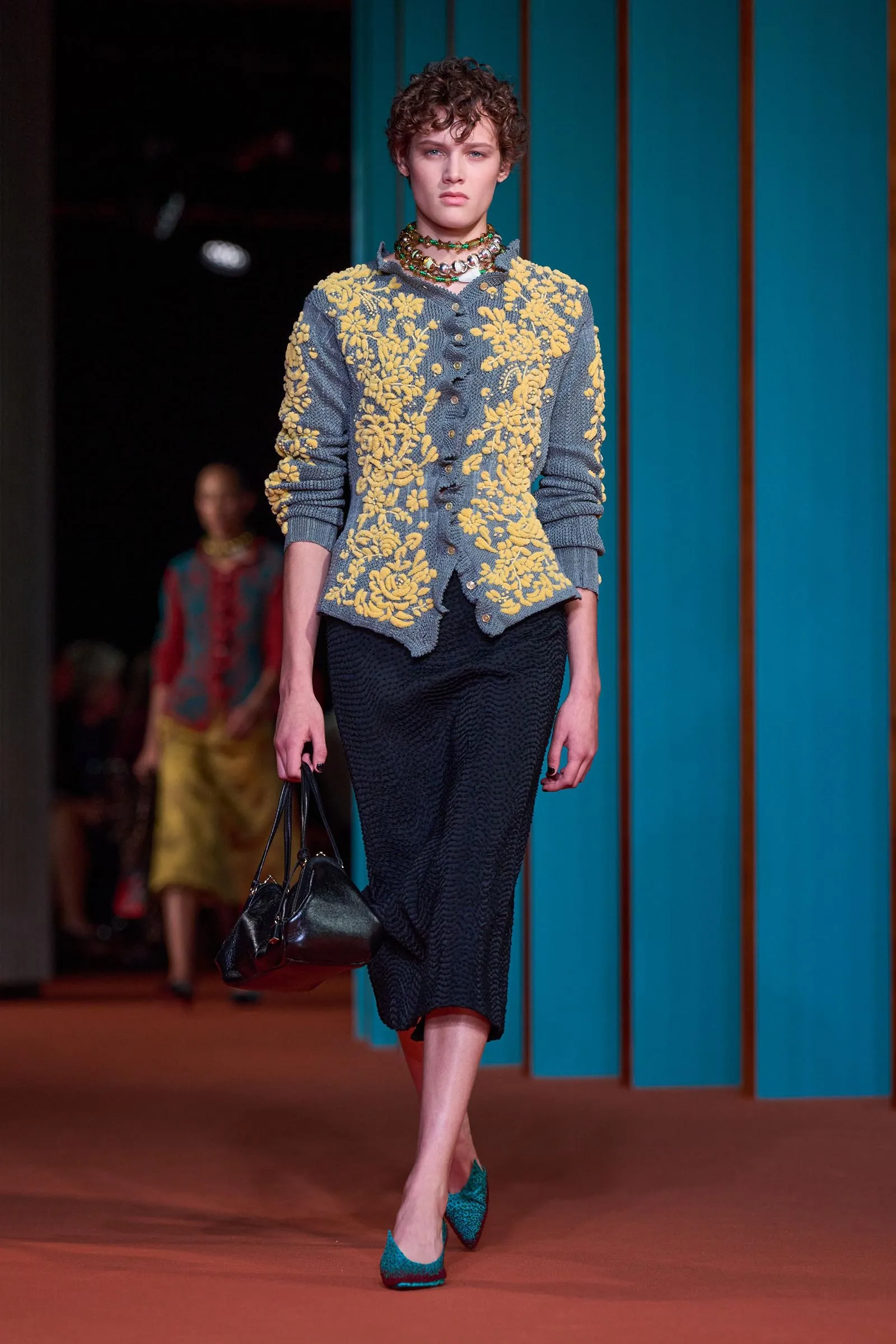 Model on a runway wearing a blue cardigan with yellow floral embroidery, a dark textured skirt, turquoise heels, layered necklaces, and a black handbag in front of teal panels.