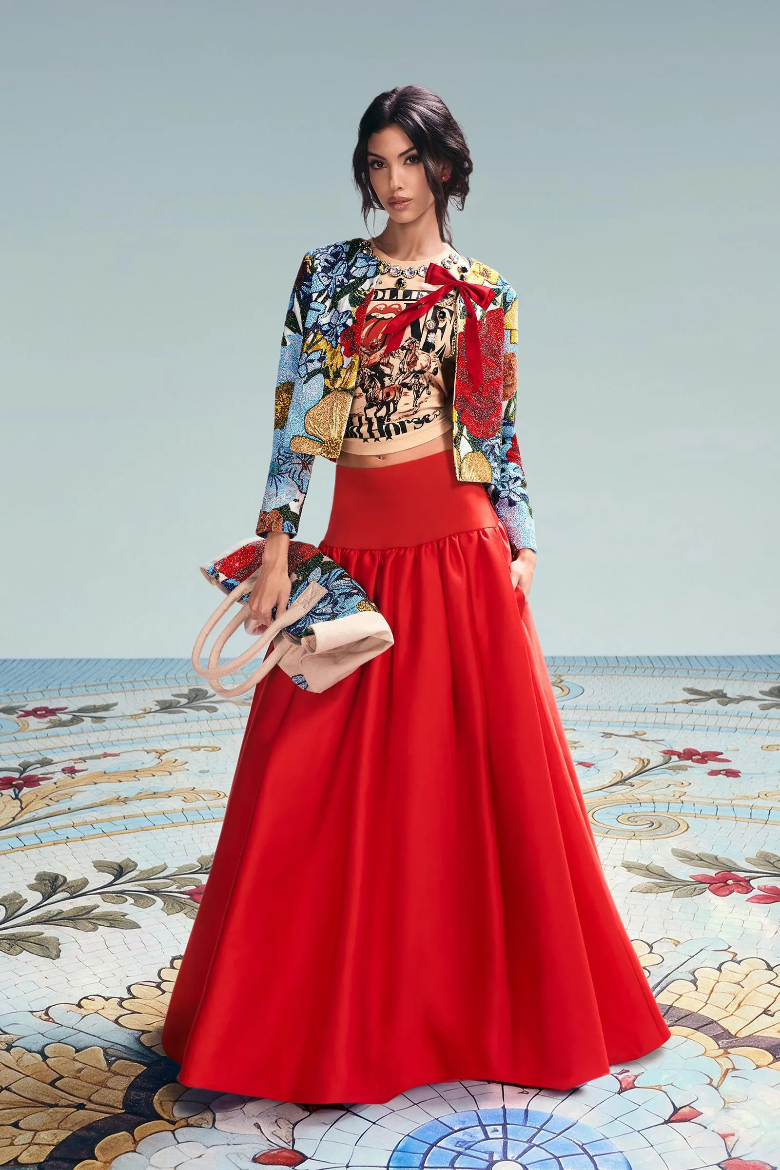 Woman wearing a colorful floral jacket and red maxi skirt, holding a patterned bag, standing on a decorative mosaic floor against a pale blue backdrop.