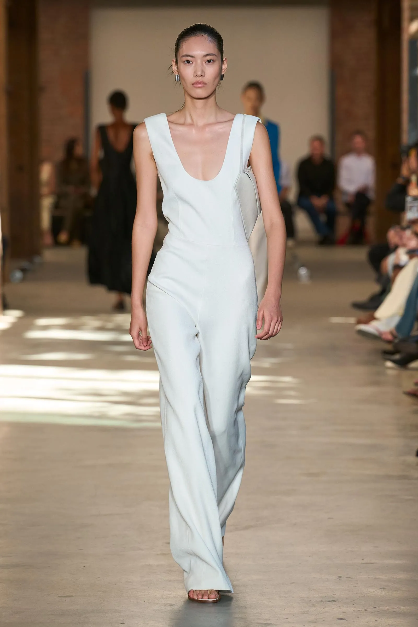 Model walking the runway in a light blue sleeveless jumpsuit with a deep V neckline, carrying a pale crossbody bag.