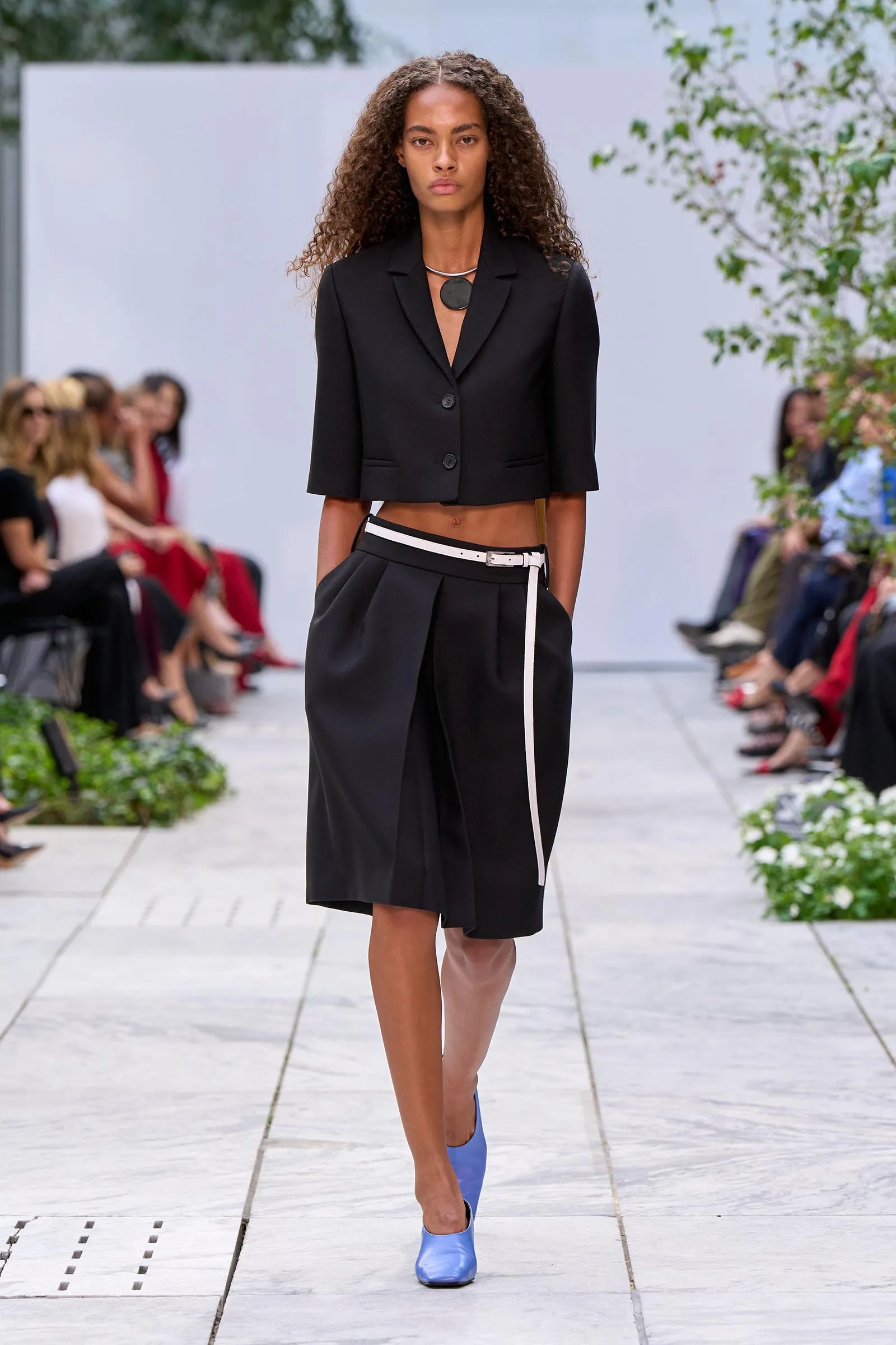 Model on an outdoor runway wearing a black cropped blazer and matching high-waisted shorts with a white belt and blue shoes.