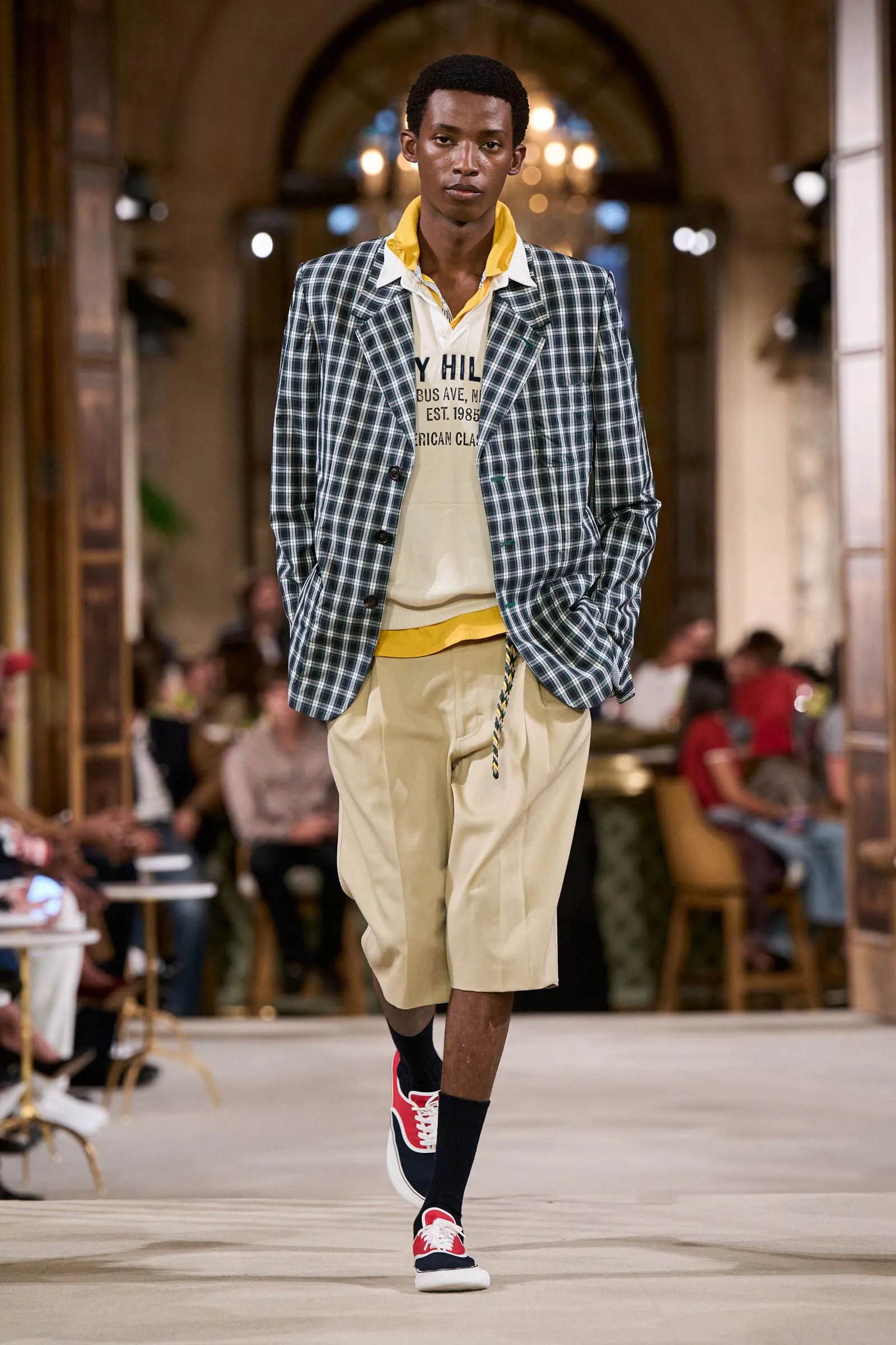 Male model on a runway wearing a navy plaid blazer, yellow shirt collar, and beige shorts, with red sneakers visible.