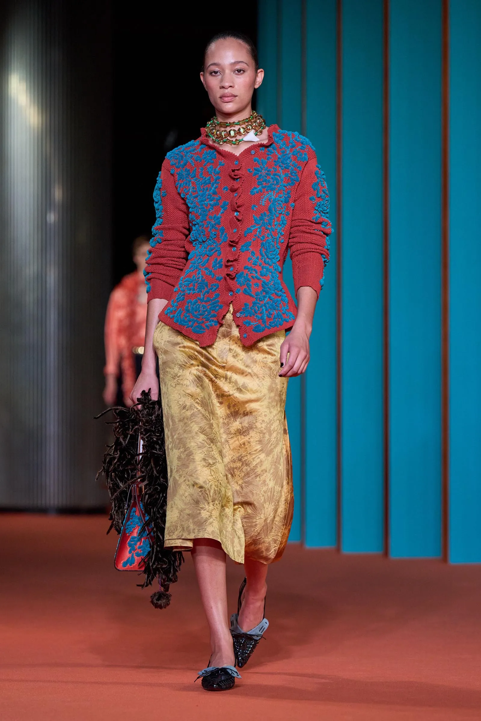 Model walking the runway in a red knit cardigan with blue floral embroidery and a gold satin skirt, against a teal backdrop.