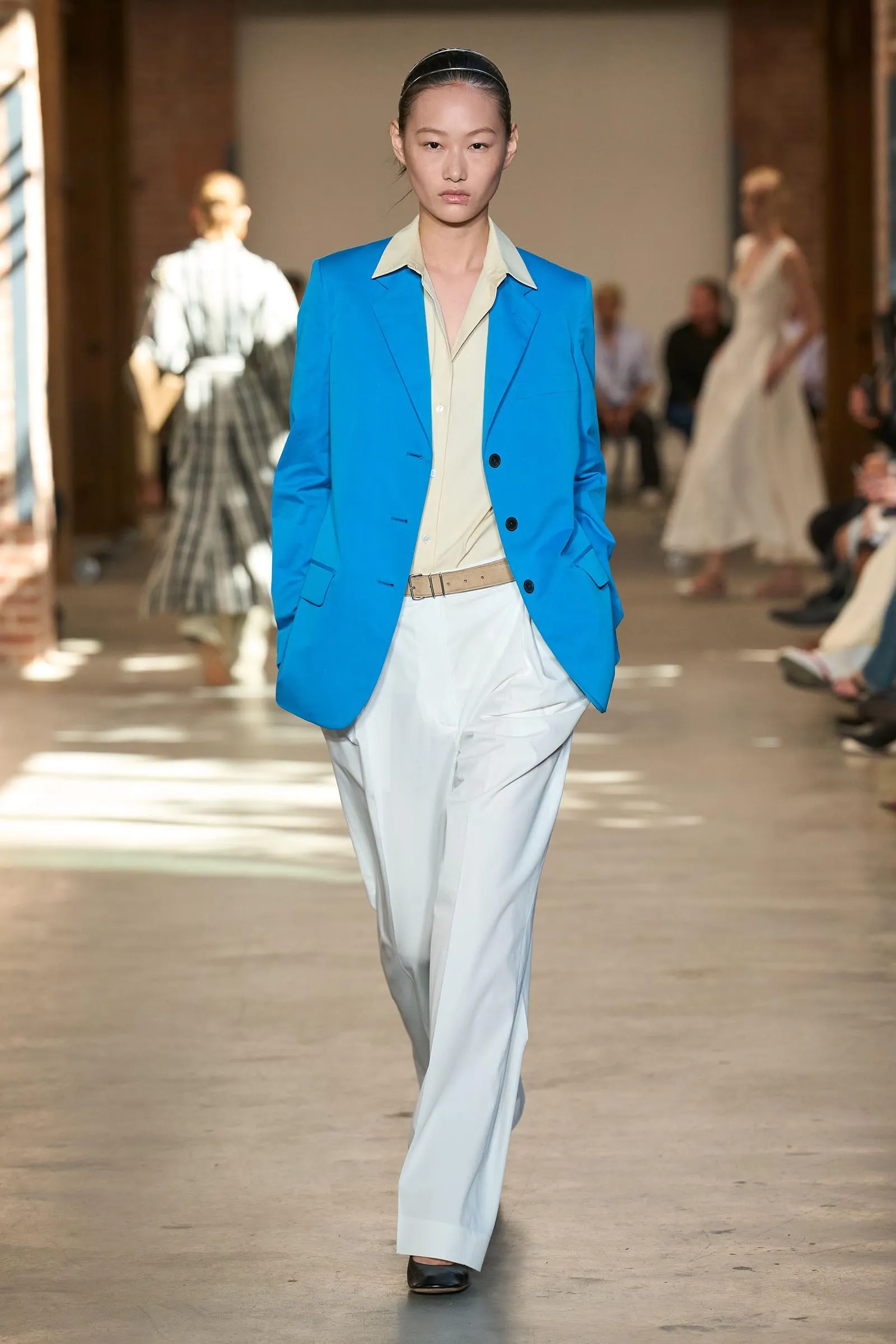 Model walks the runway wearing a bright blue blazer over a pale beige shirt and light gray trousers, accessorized with a tan belt and black shoes.