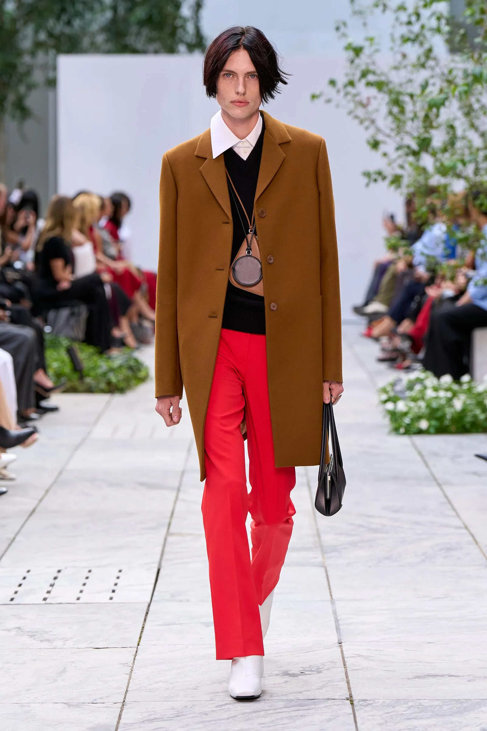 Fashion model on a runway wearing a camel coat over a black outfit, bright red trousers, white boots, and carrying a black handbag.