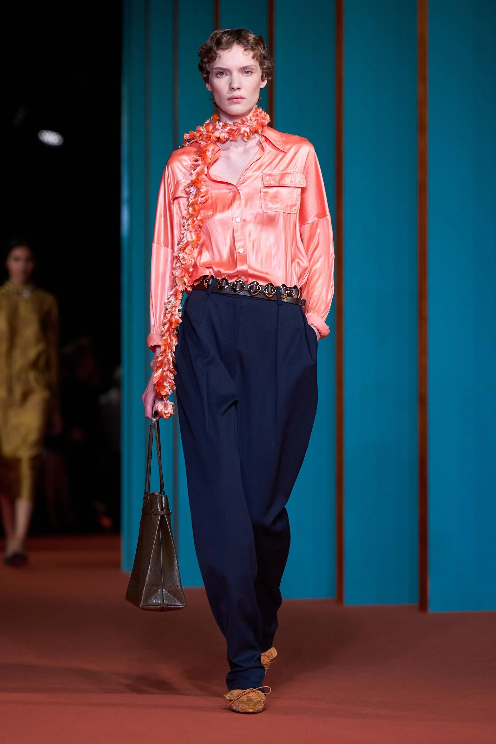 Runway model wearing coral satin shirt with navy pants, an orange flower lei, and a brown handbag against a teal backdrop.