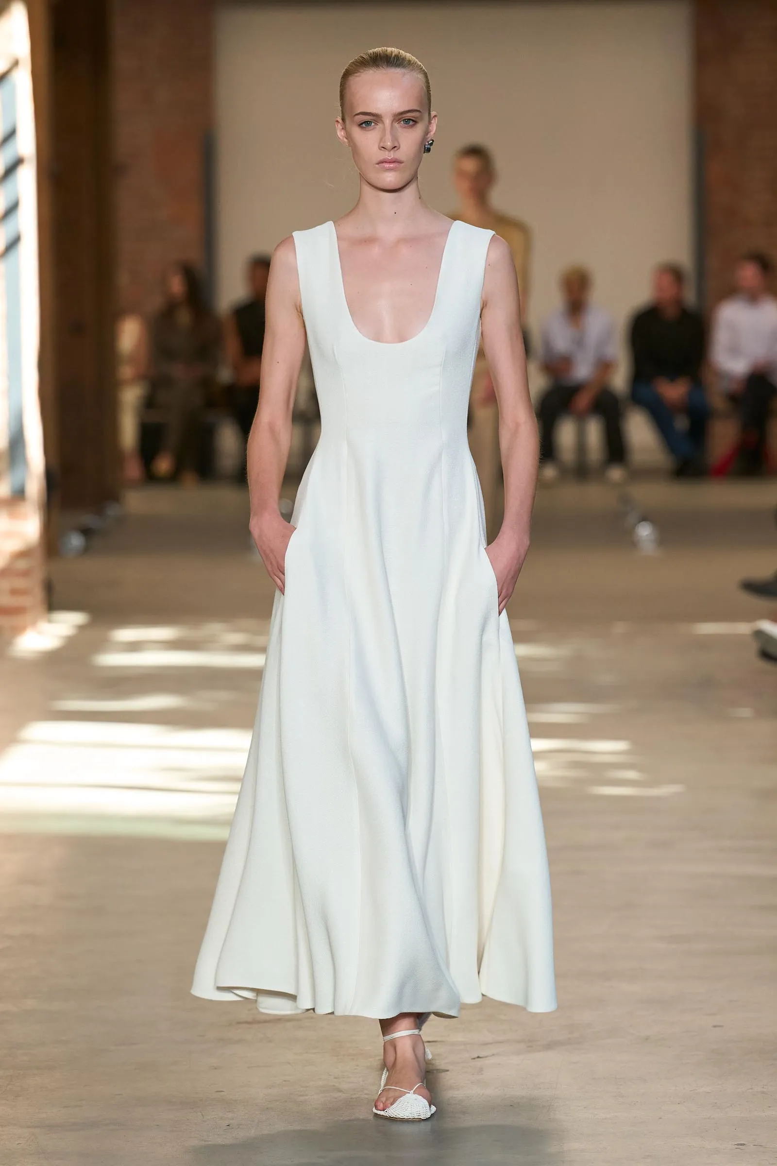 Model walking a runway in a white sleeveless dress with a deep scoop neckline and side pockets, audience blurred behind.