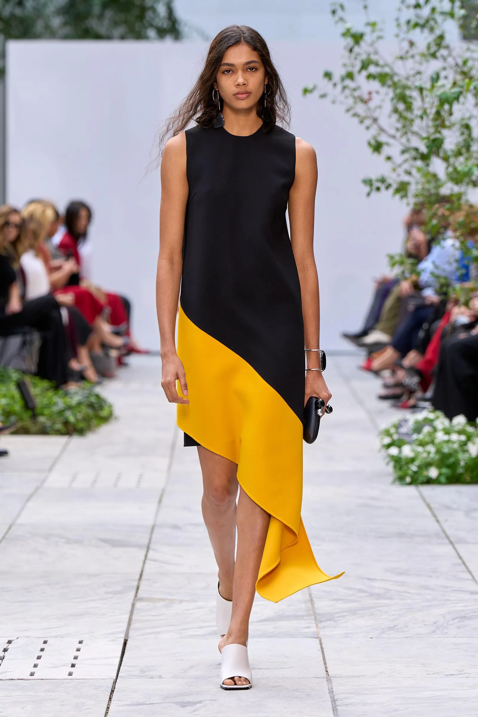 Model on a runway wearing a sleeveless black and yellow diagonal color-block dress with white sandals, holding a black clutch.