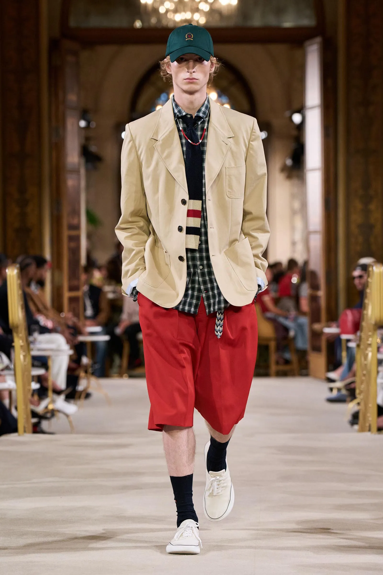 Male model walks the runway in a beige blazer, plaid shirt, red shorts, and a green cap in a grand indoor setting.