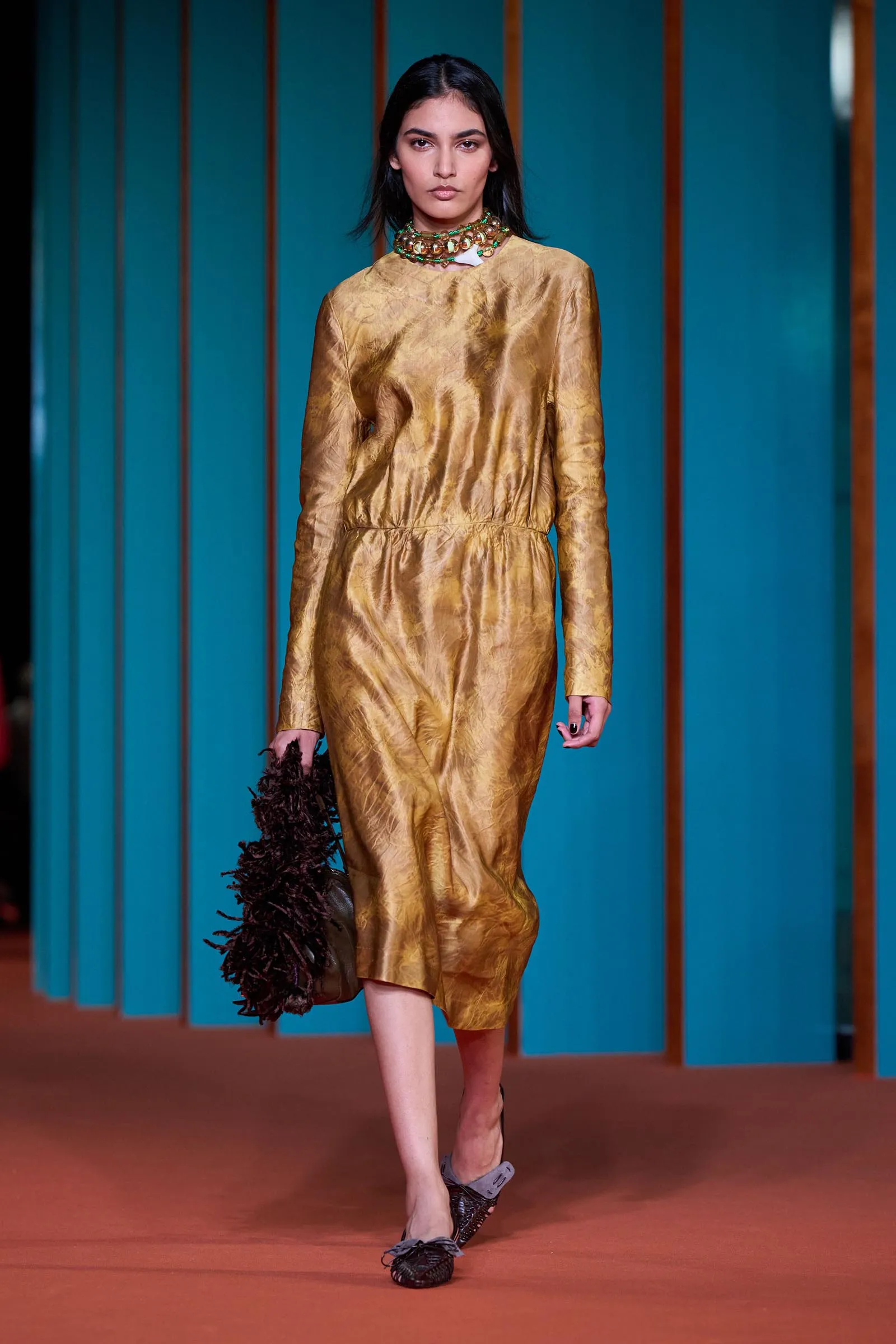 Model walking runway in a gold long-sleeve dress with emerald necklace against teal vertical panels.