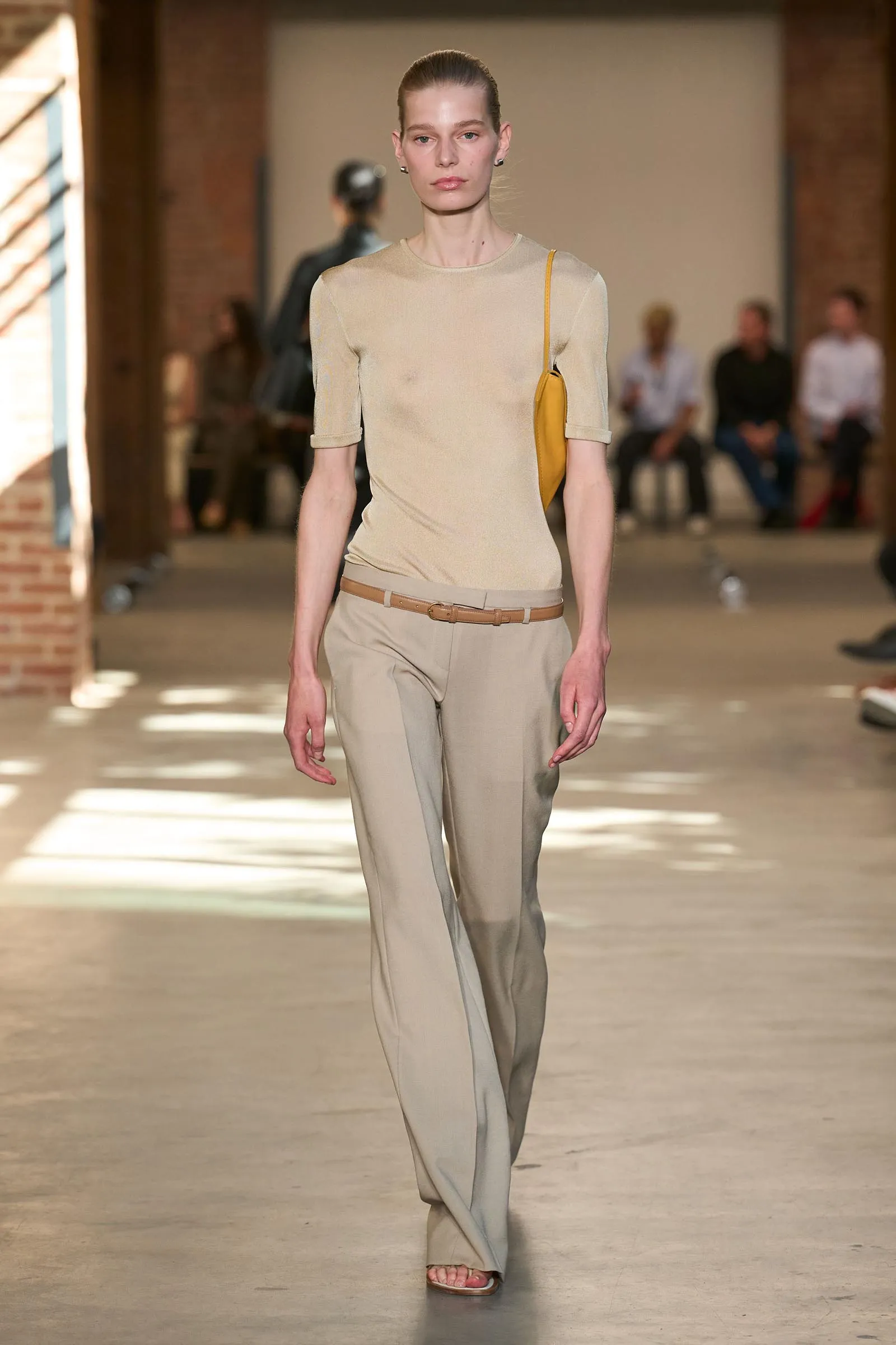 Model walks down a runway in beige top and matching wide-leg pants, yellow shoulder bag, neutral-toned belt.
