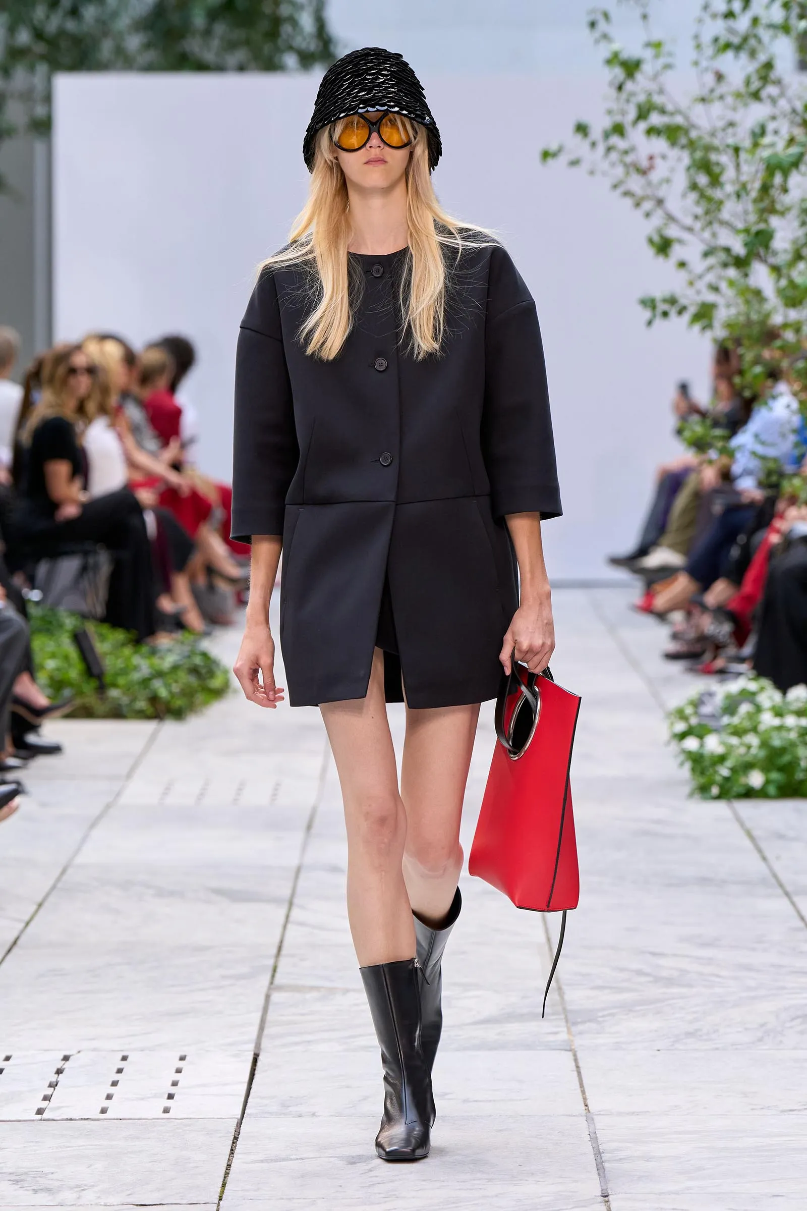 Model walks the runway in a black coat with a matching hat, orange sunglasses, knee-high boots, and a red tote bag on the side.