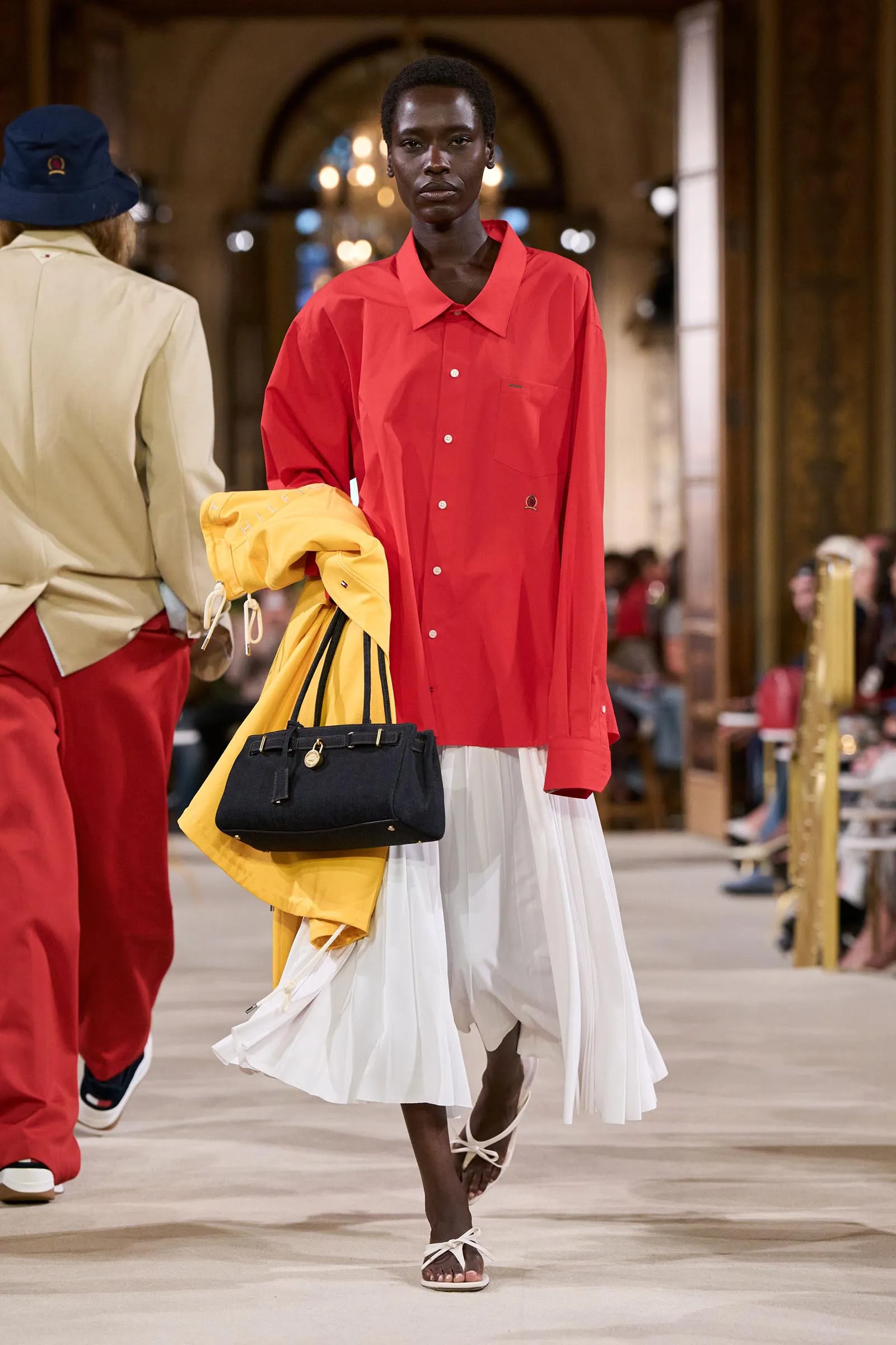 Model walks a runway in a bright red button-down shirt and white pleated skirt, carrying a yellow jacket and a black handbag.