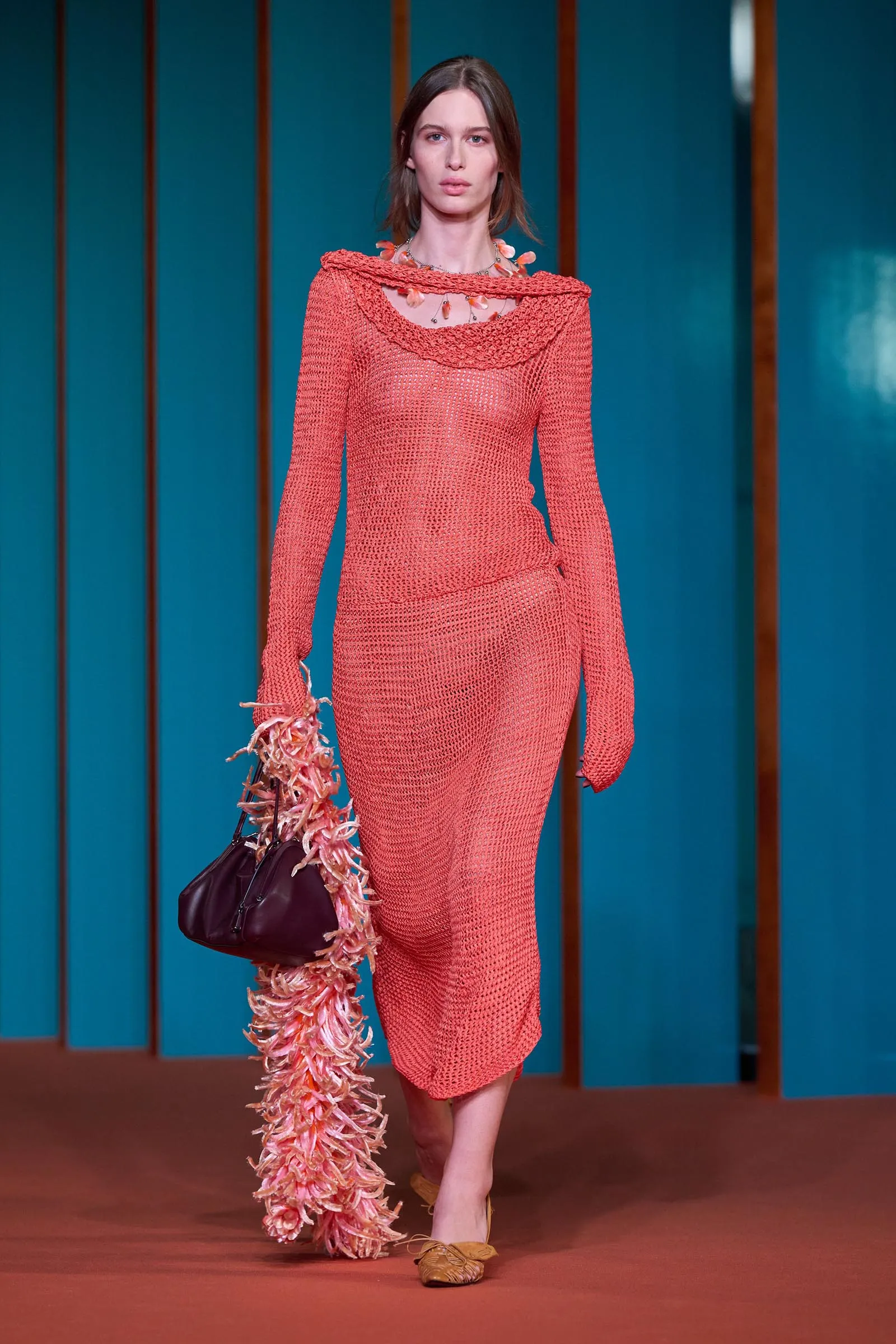 Model walking a fashion show in a coral knit dress, holding a black handbag with a pink feather scarf, teal panel backdrop.