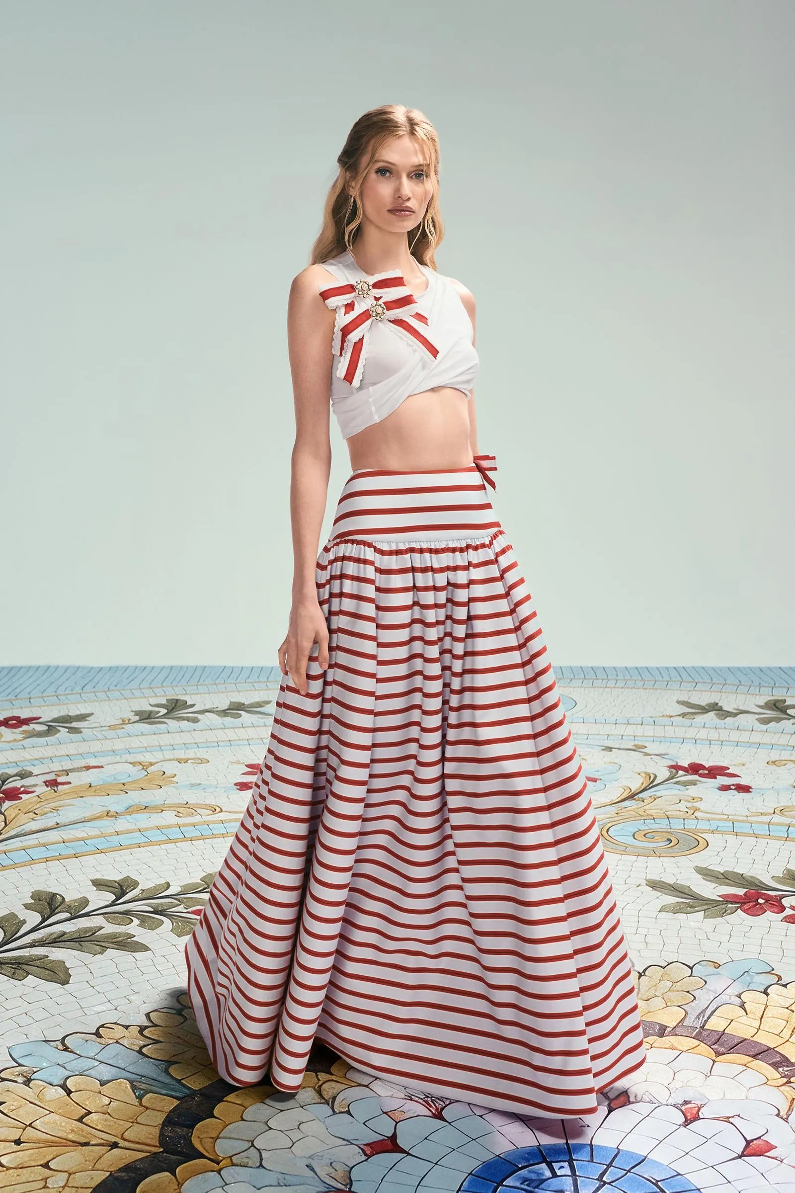 Model wearing a white cropped top with red ribbon accents and a red-and-white striped high-waisted skirt, standing on a decorative tiled floor.