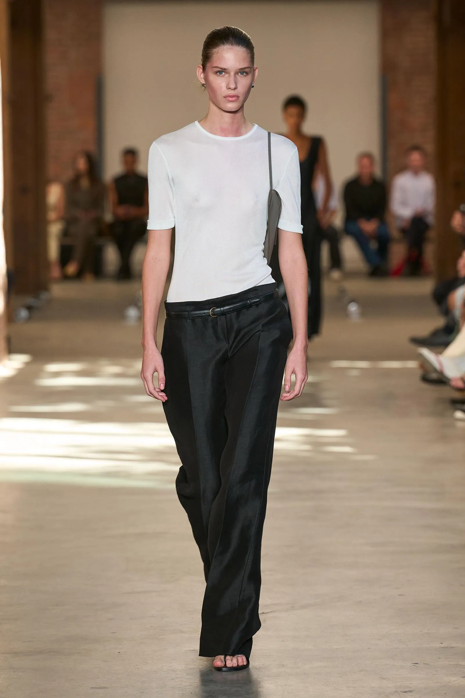 Model walking a fashion runway in a white fitted tee, black wide-leg pants, and a gray shoulder bag, with onlookers in the background.