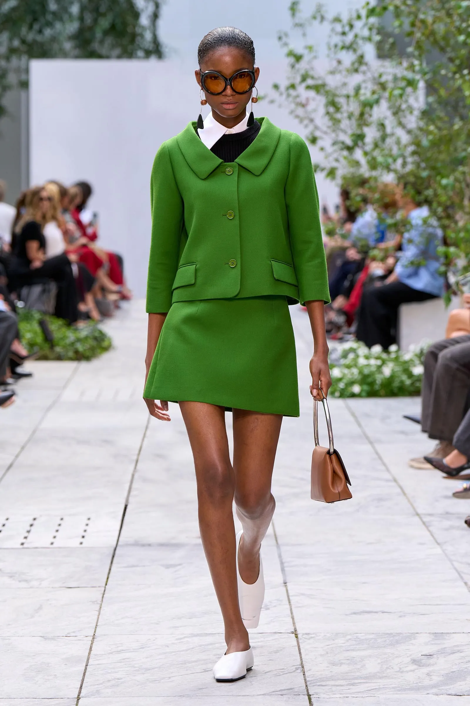 Model on a fashion runway wearing a bright green tailored jacket with matching skirt, white shoes, oversized sunglasses, and a tan handbag.