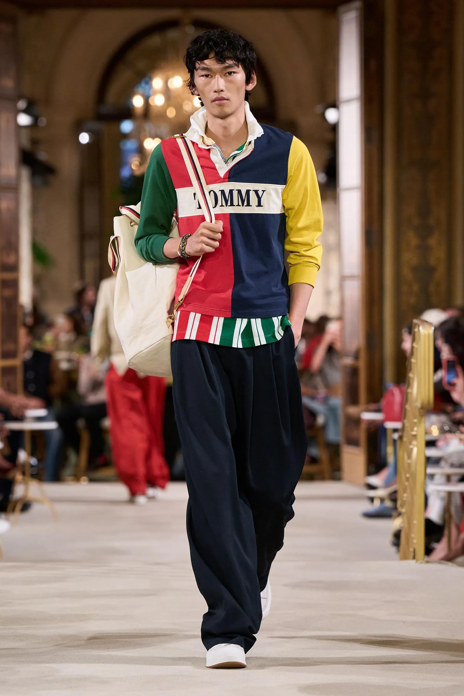 Male model walks a runway in a color-blocked shirt with 'TOMMY', navy pants, white sneakers, and a beige tote bag over his shoulder.