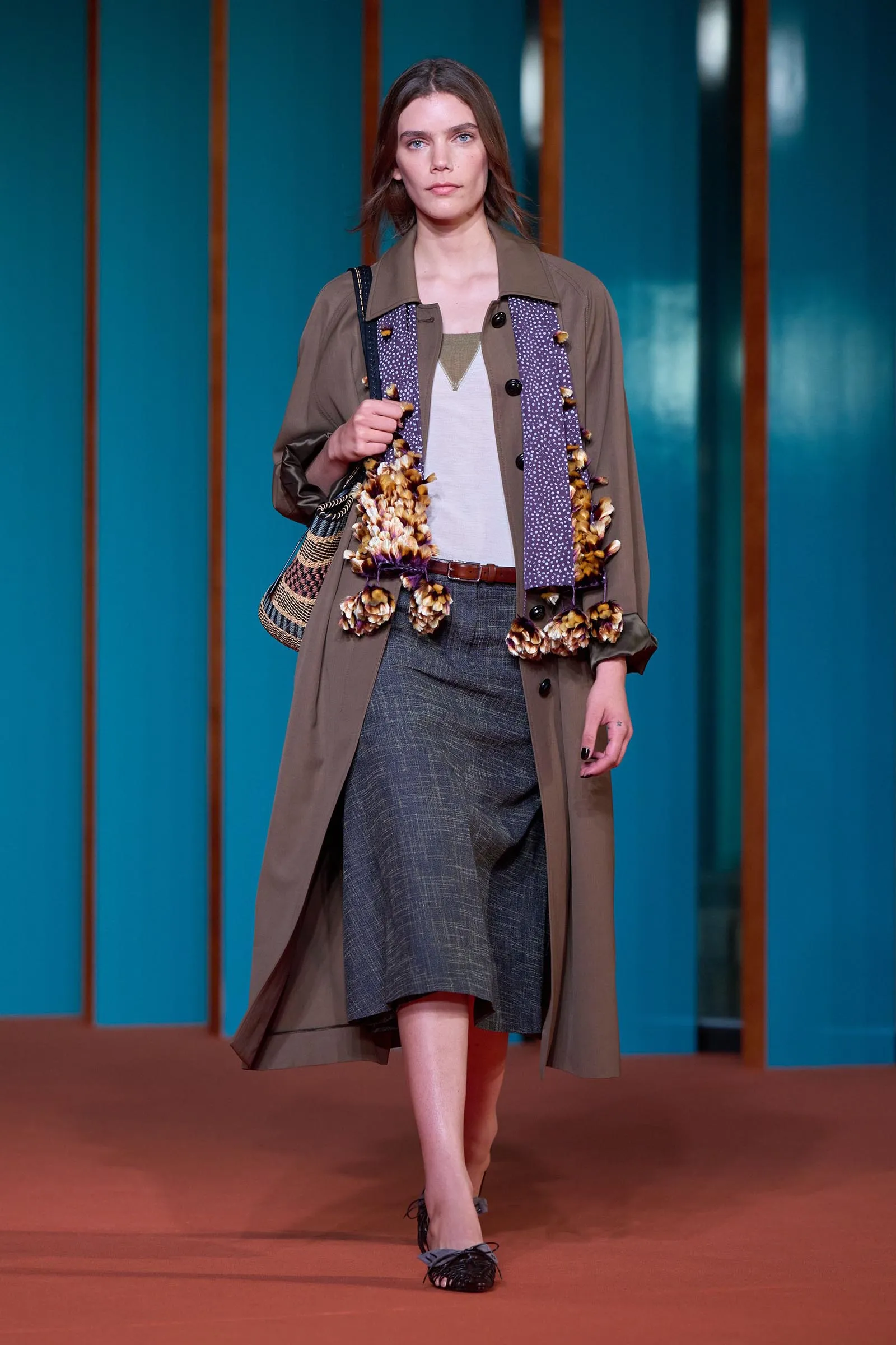 Runway model walking in a tan trench over a white top and gray plaid skirt, with a purple patterned scarf and a woven shoulder bag.