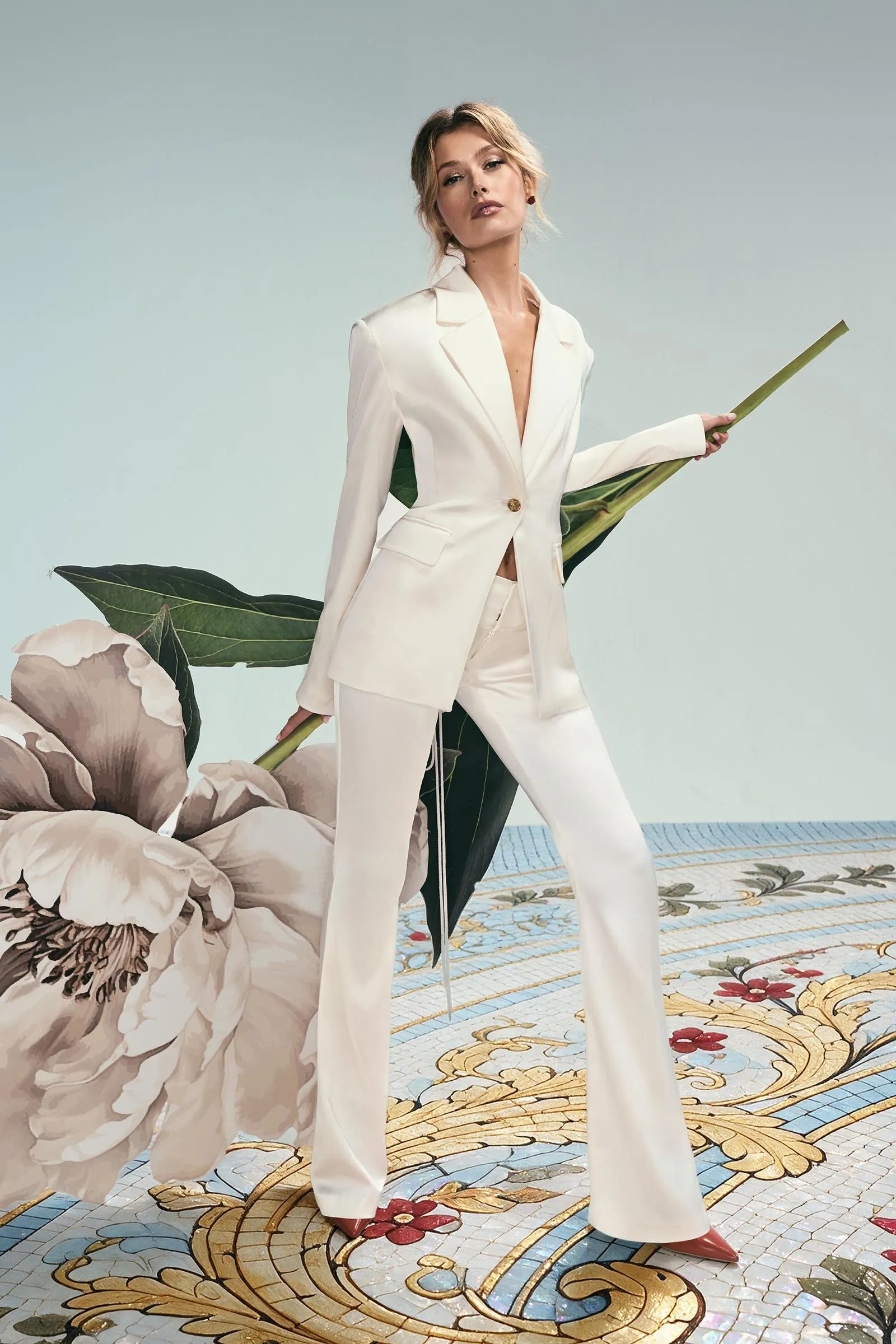 Model in a white suit holding a long green leaf, posing on a floral tiled floor with oversized blossoms nearby.