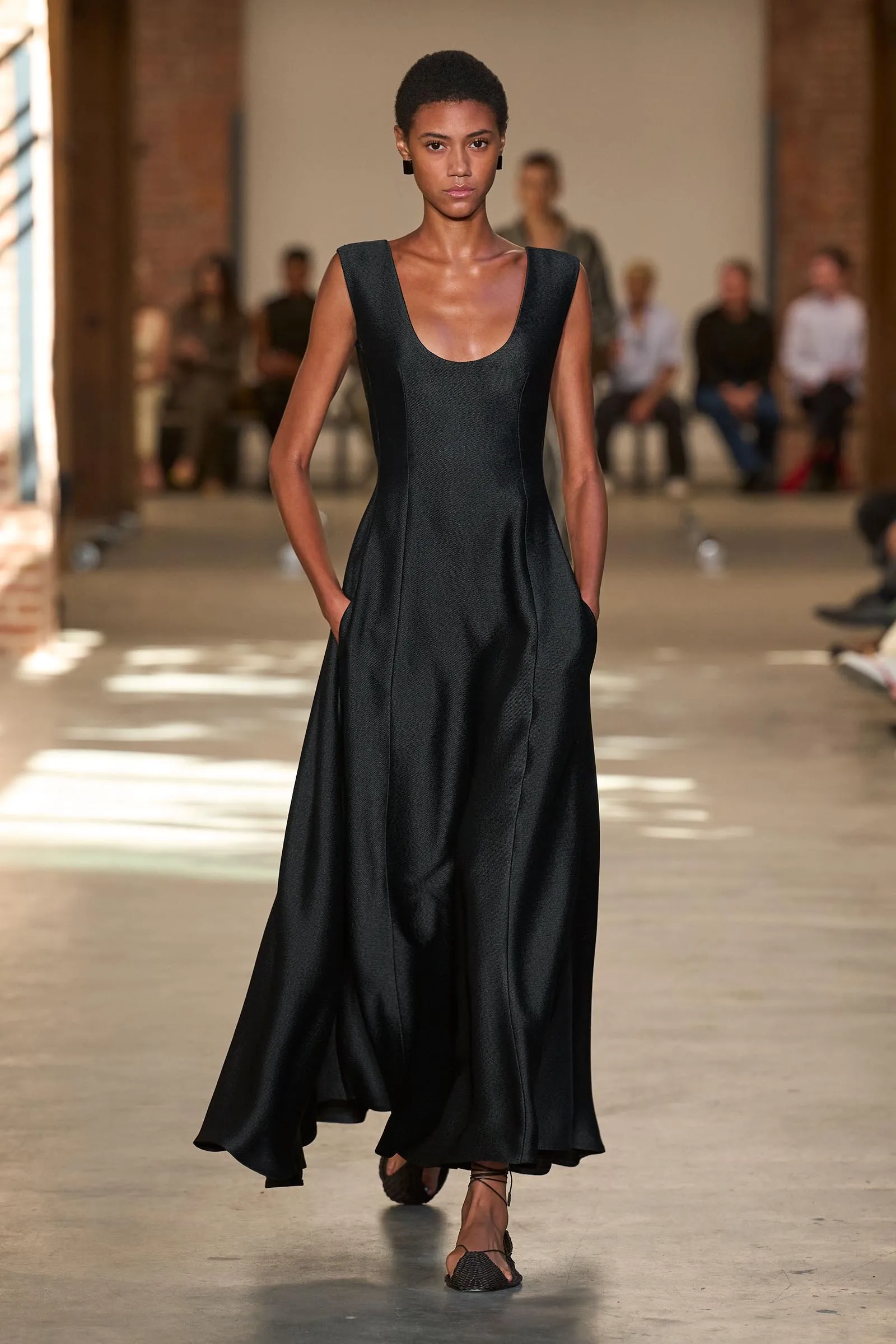 Model walking down a runway in a sleeveless black evening gown with pockets, audience blurred in the background.