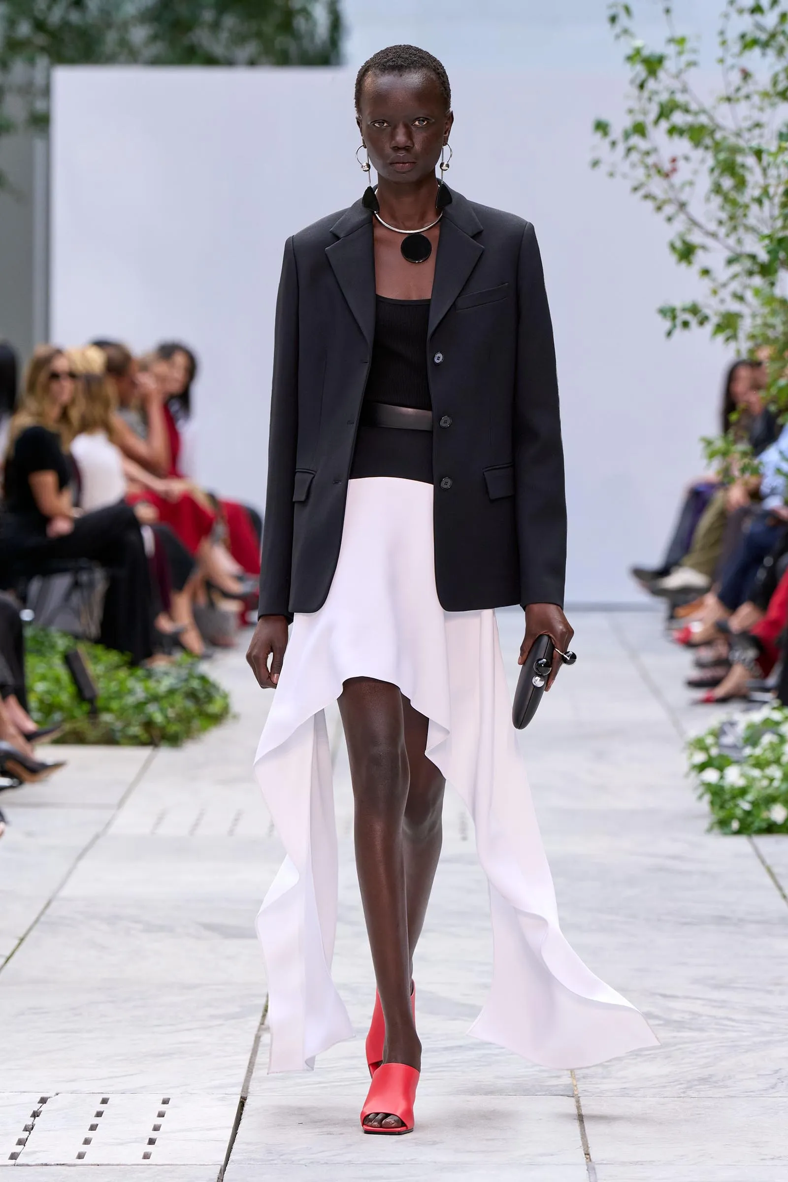 Model on a runway wearing a black tailored blazer over a black top, white high-low skirt, and bright red sandals, with bold jewelry.