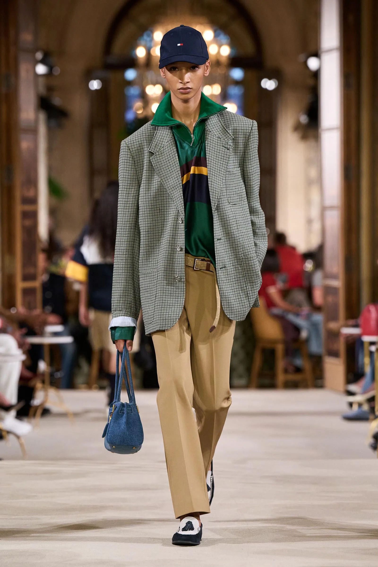 Model walking a fashion runway in a gray check blazer, green polo, tan trousers, blue bag, and navy cap in a bright indoor venue.