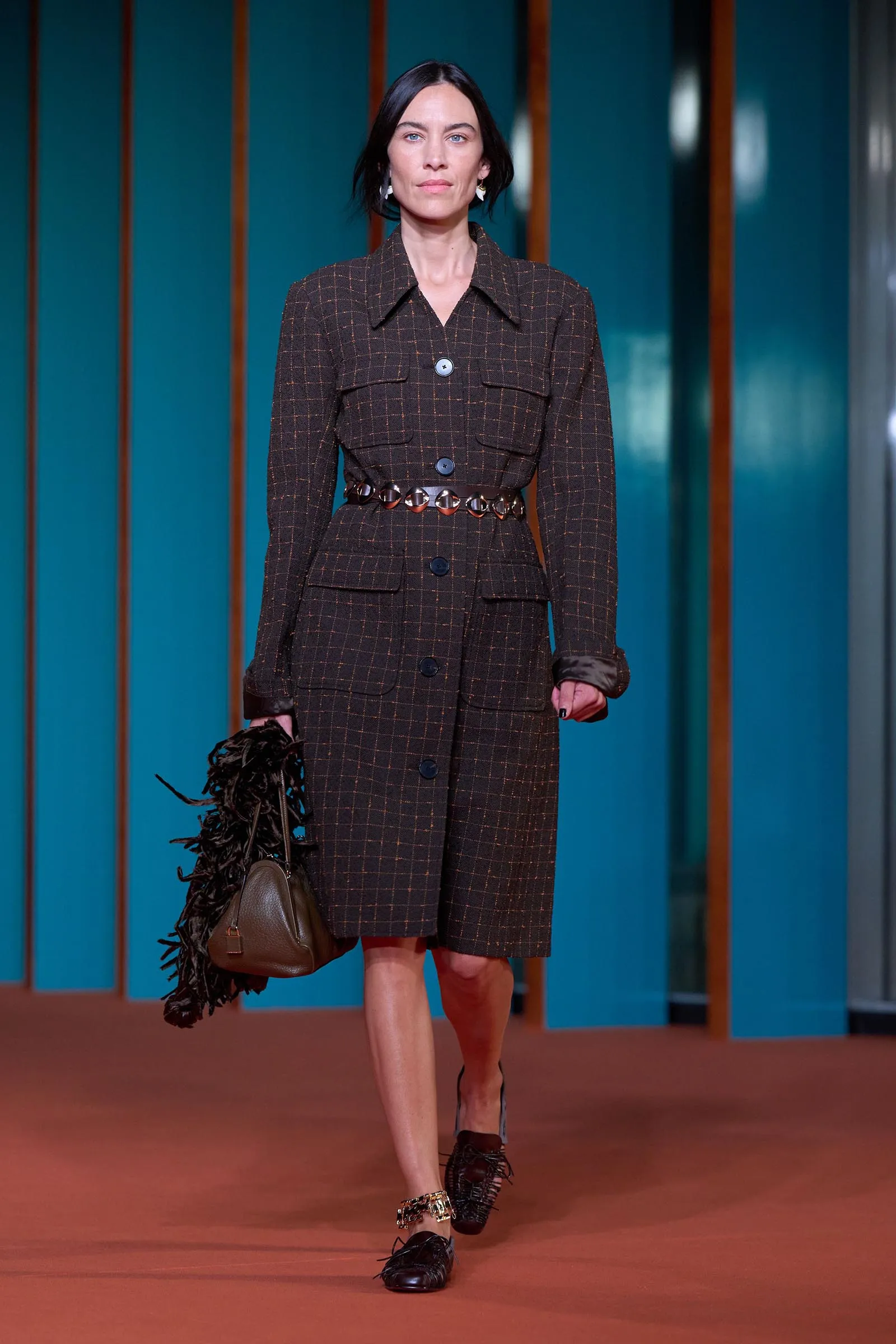 Runway model wearing a dark brown plaid belted dress with large buttons and pockets, carrying a brown handbag with a feathered accessory against teal backdrop.