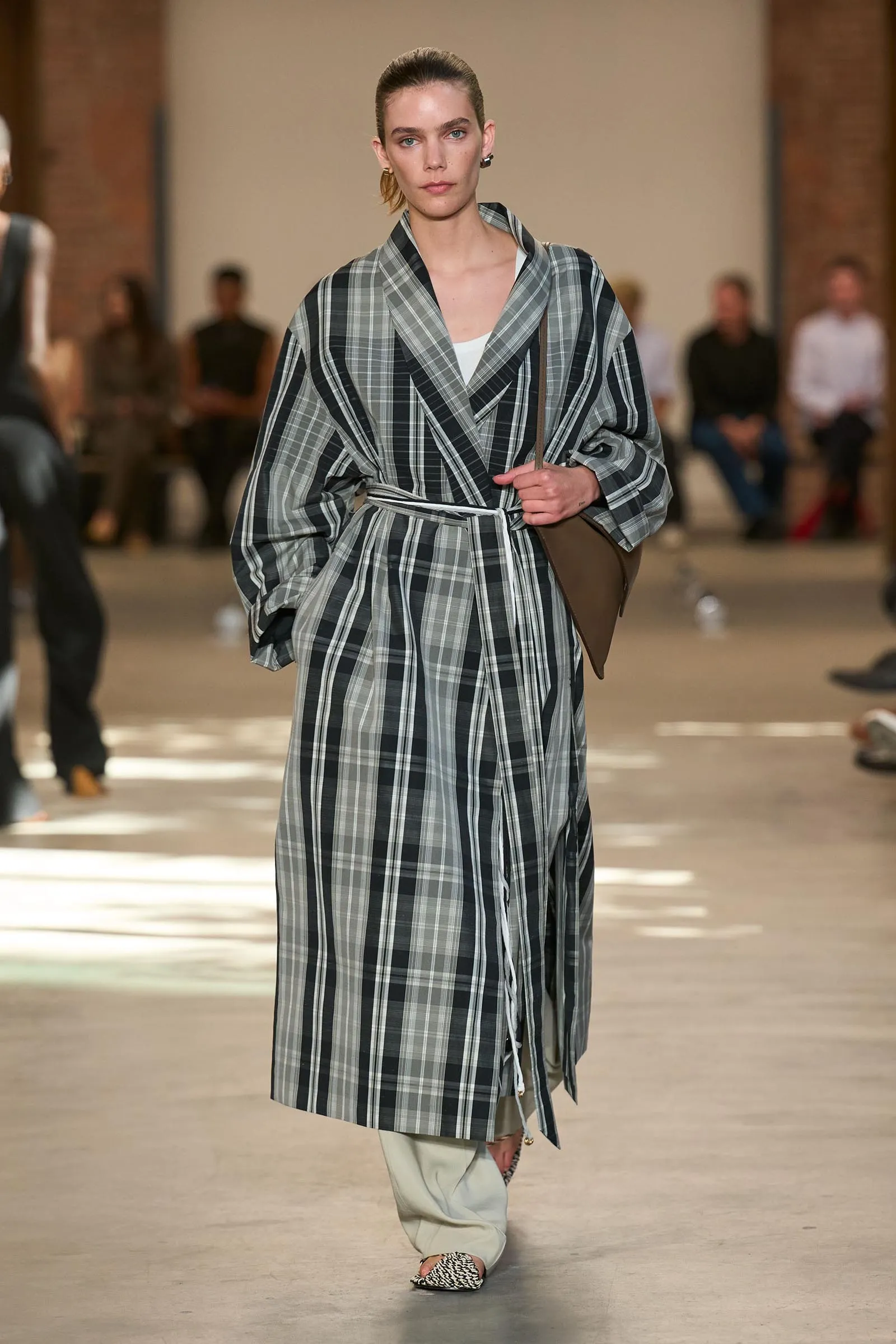 Model struts down a runway in a gray plaid wrap coat with a belt, white top, and light trousers; leopard-print flats complete the look.