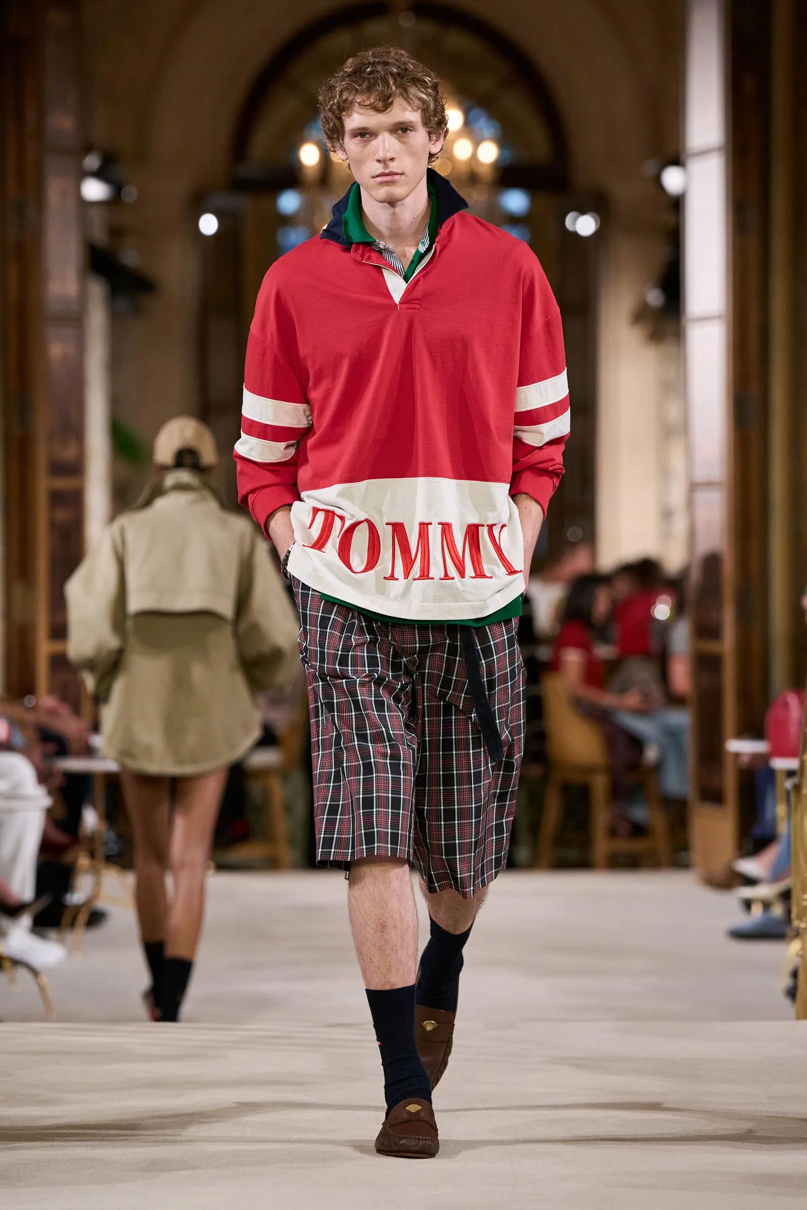 Male model walking a runway in a red long-sleeve polo with white accents and a white hem band that reads 'TOMMY', paired with plaid shorts in a fashion show setting.