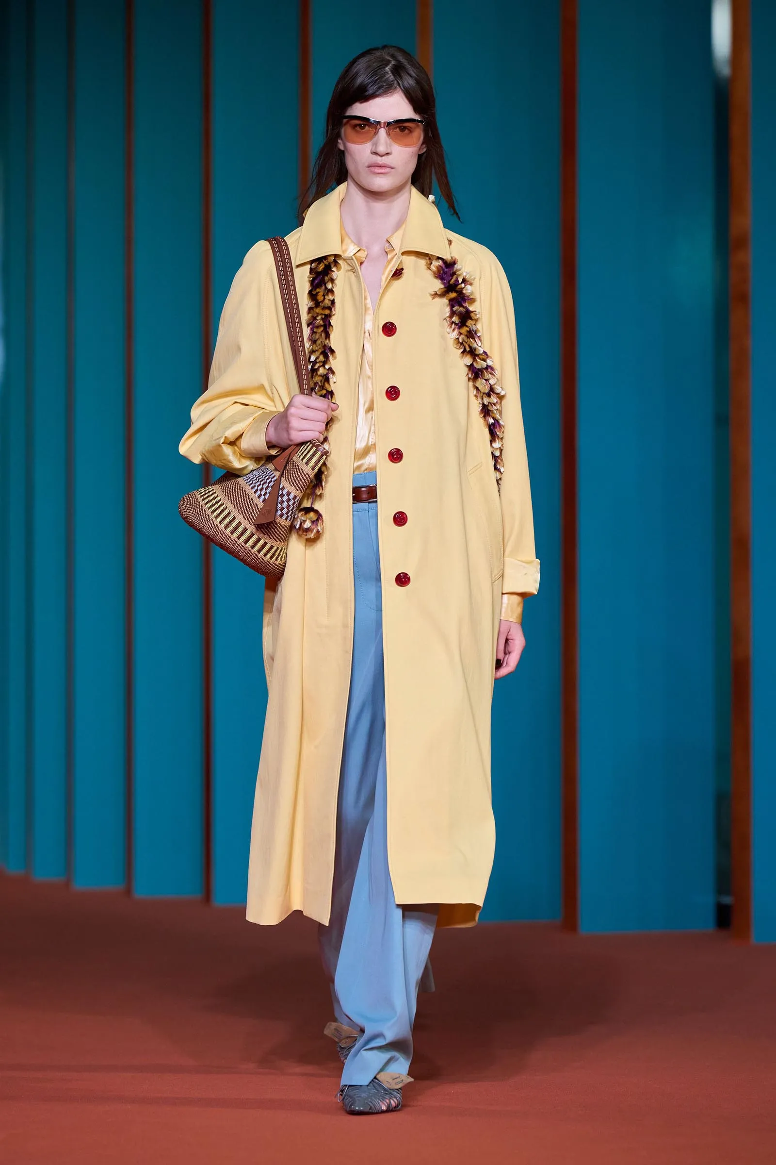 Model walking a fashion runway in a yellow trench coat over pale blue pants, orange sunglasses, and a woven brown bag with a floral garland draped over the shoulders.