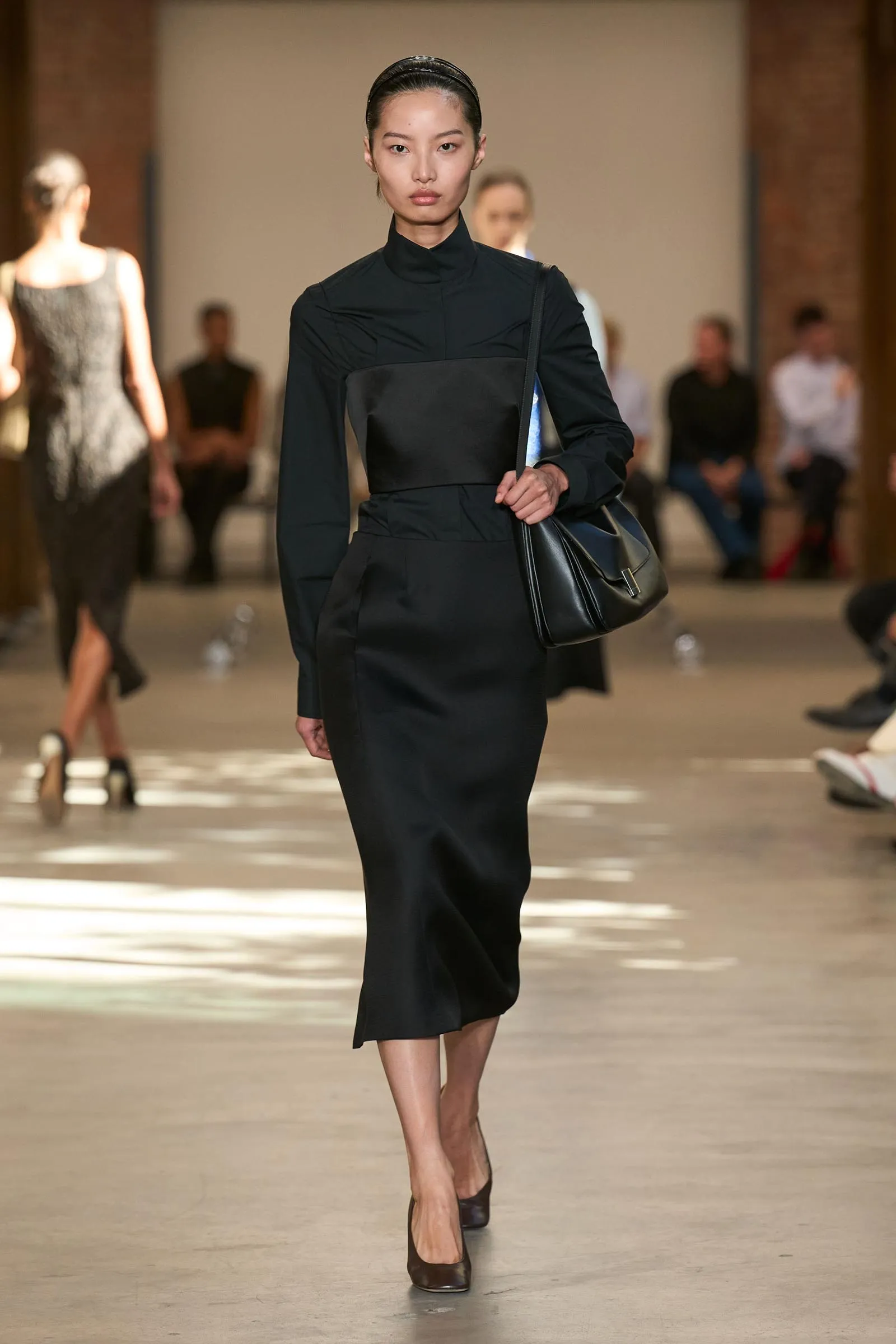 Model walking a runway in a black turtleneck dress, carrying a black leather shoulder bag, with a glossy headband and brown heels.
