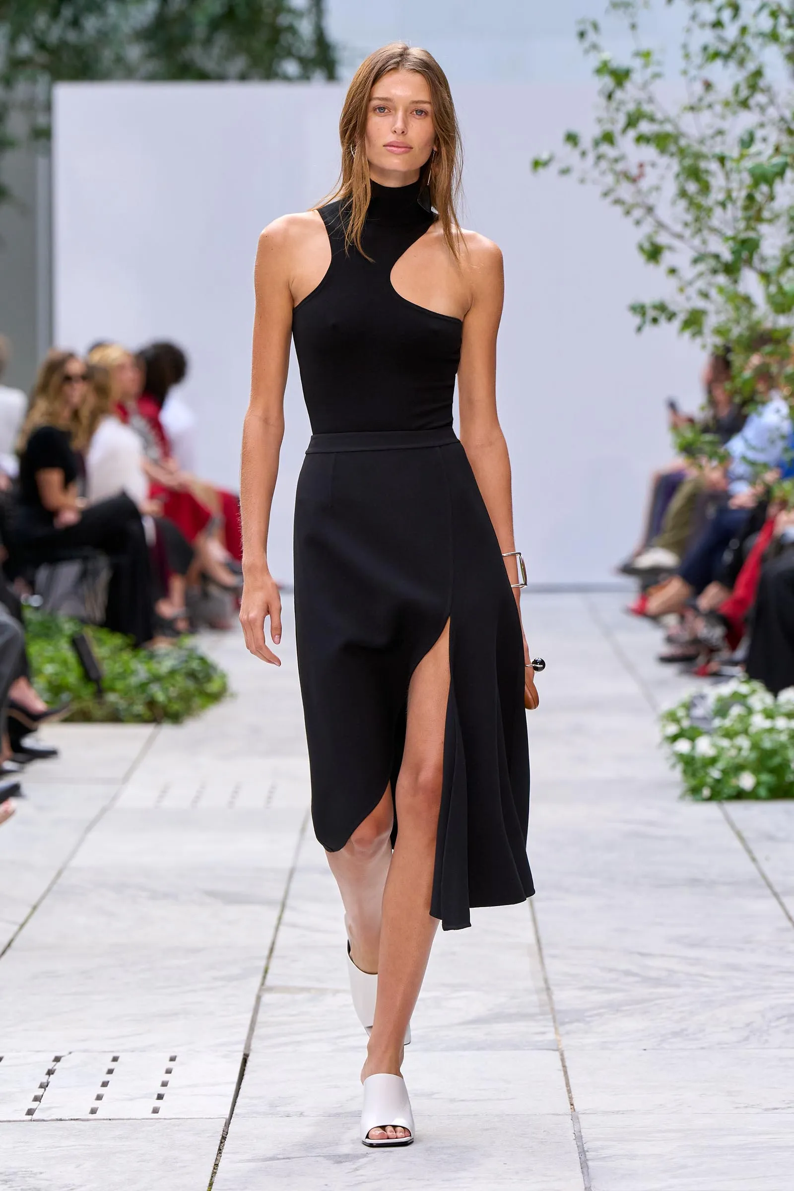 Model walks the runway in a black, asymmetrical one-shoulder dress with a thigh-high slit, white sandals, outdoor show with spectators in the background.