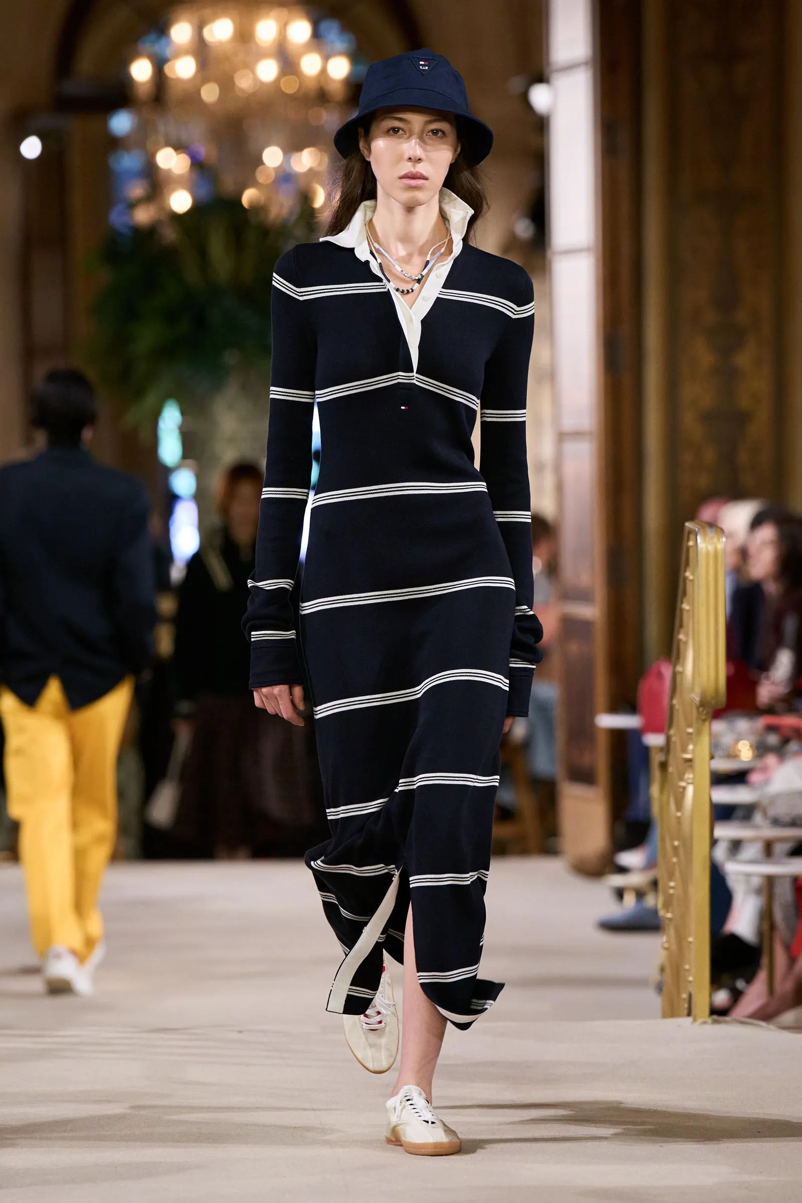 Fashion model walking a runway in a navy striped dress with white sneakers and a blue bucket hat in a grand indoor venue.