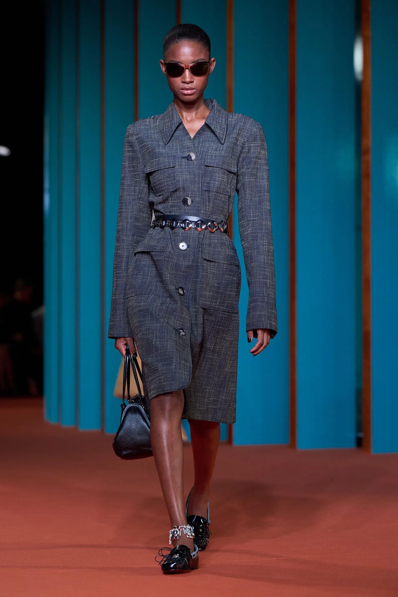 Model walks the runway in a gray plaid blazer-dress with a matching belt and black accessories, teal striped backdrop behind.