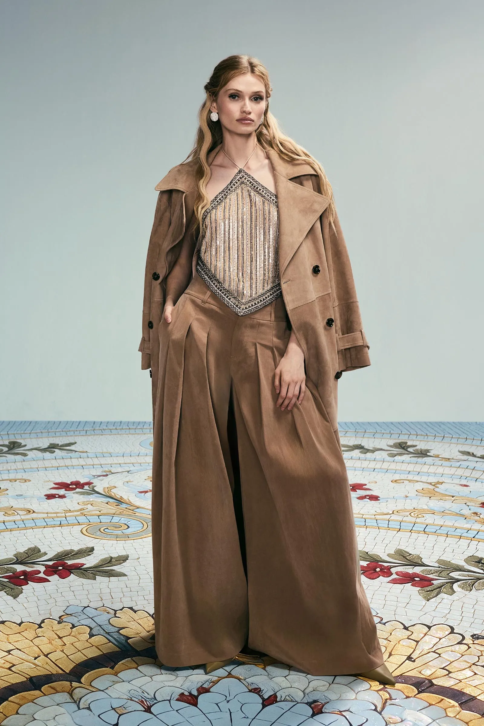 Fashionable woman in a tan trench coat over a beaded top, posing against a pale blue backdrop on a patterned tile floor.