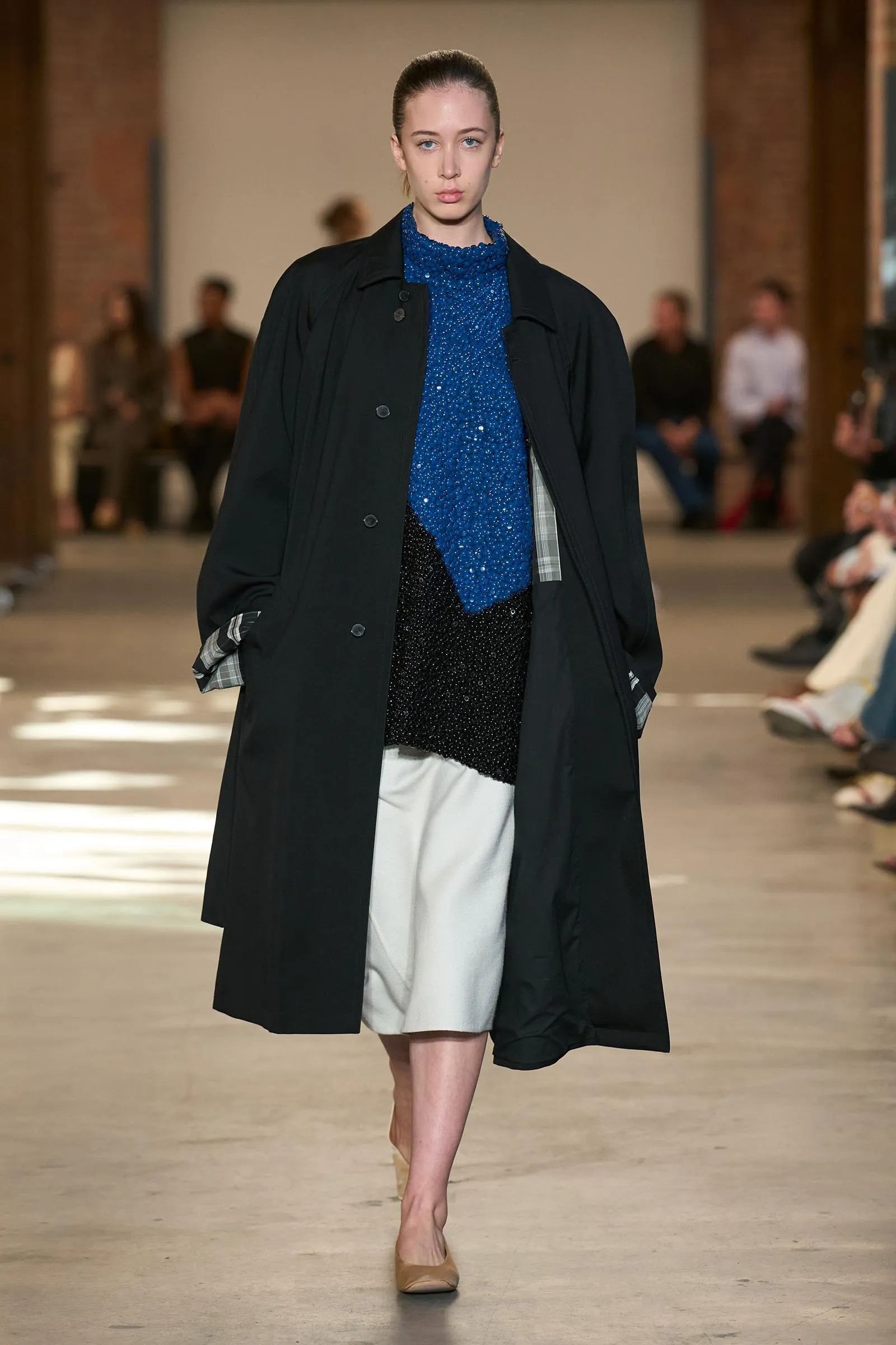 Model walking a runway in a blue glittery top under a long black coat and a light skirt, during a fashion show.