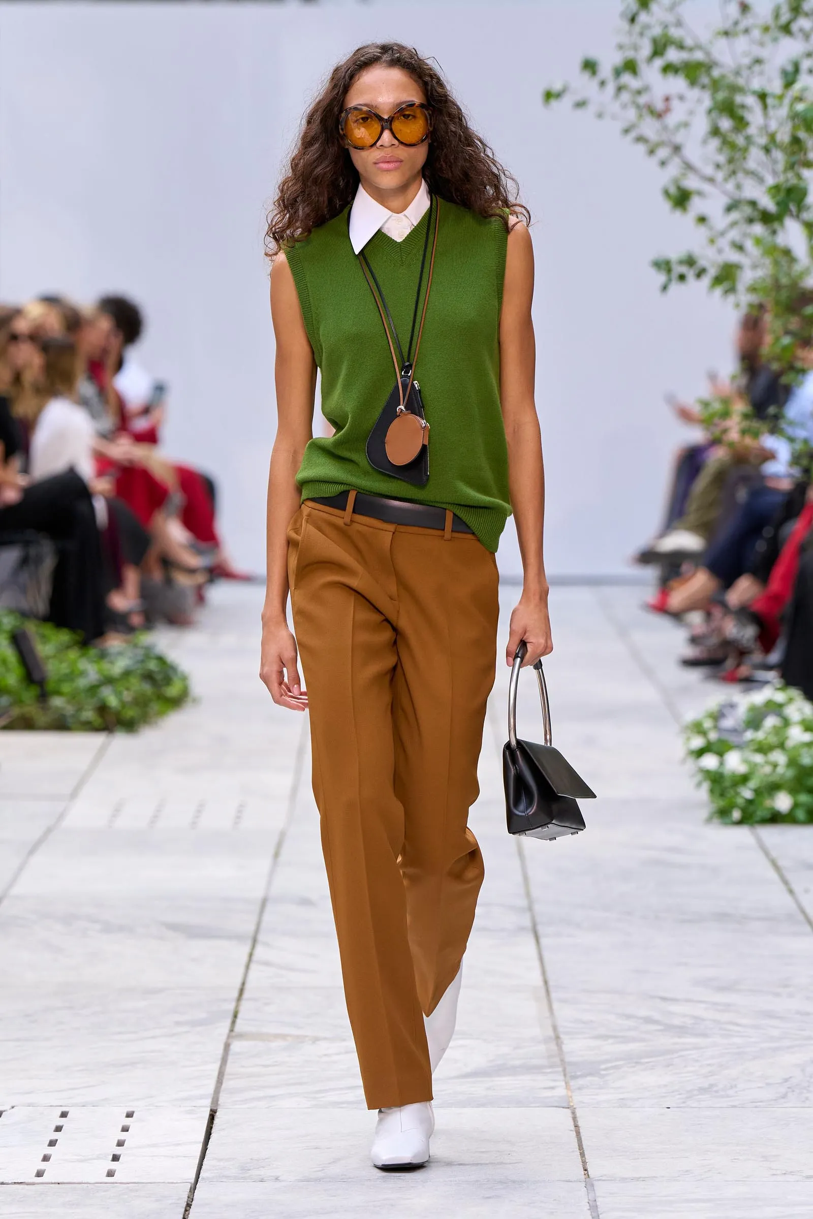 Model walking a runway in a green sleeveless knit over a white shirt, tan trousers, white boots, and oversized orange sunglasses with a black handbag accessory.